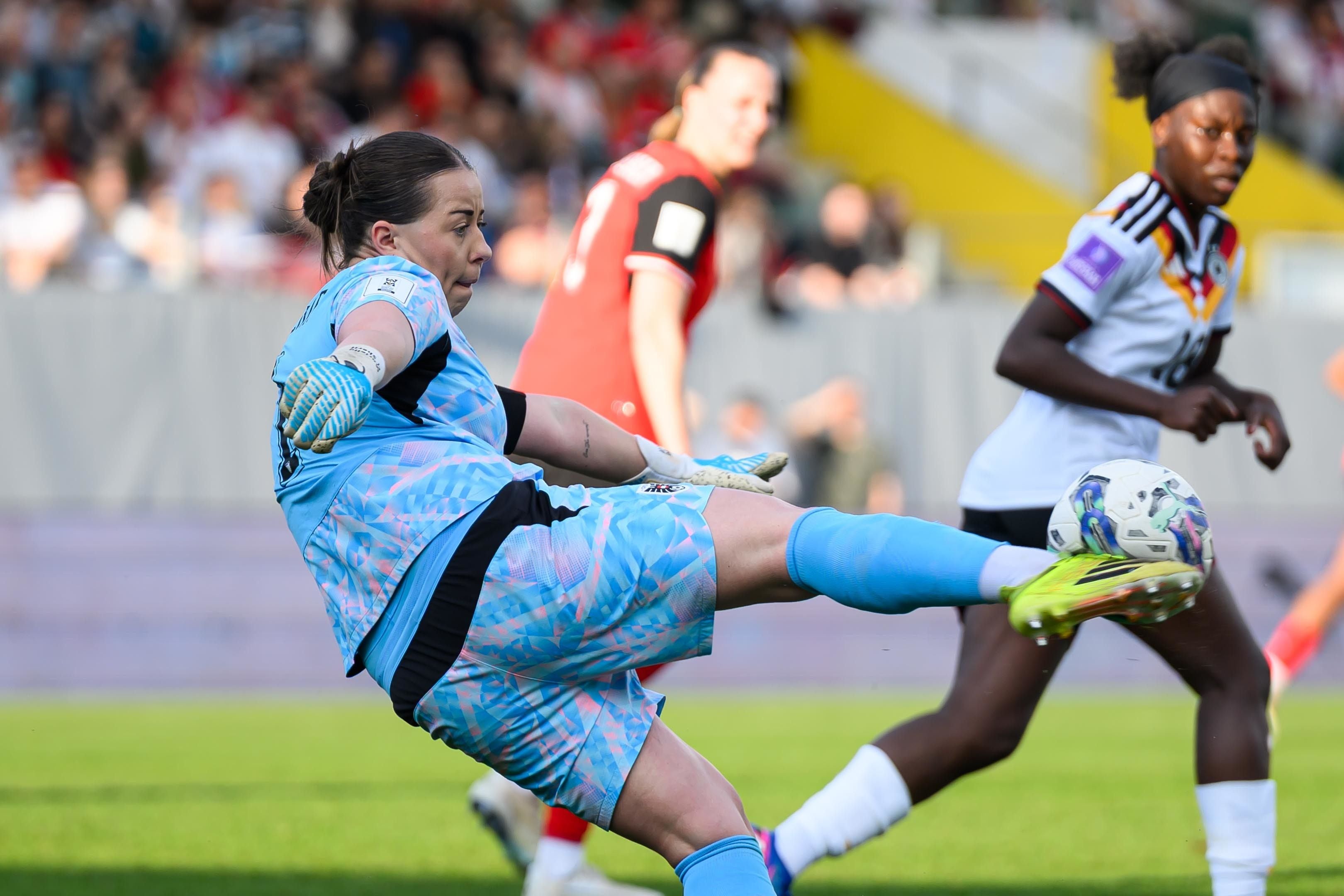 ÖFB-Torfrau Mariella El Sherif im hellblauen Trikot kickt den Fußball mit dem rechten Fuß hoch in der Luft weg, während im Hintergrund zwei weitere Spielerinnen und Zuschauer auf den Tribünen zu sehen sind. Die Österreicherinnen rettete mit starken Paraden und vollem Einsatz den knappen 1:0-Sieg gegen Slowenien.