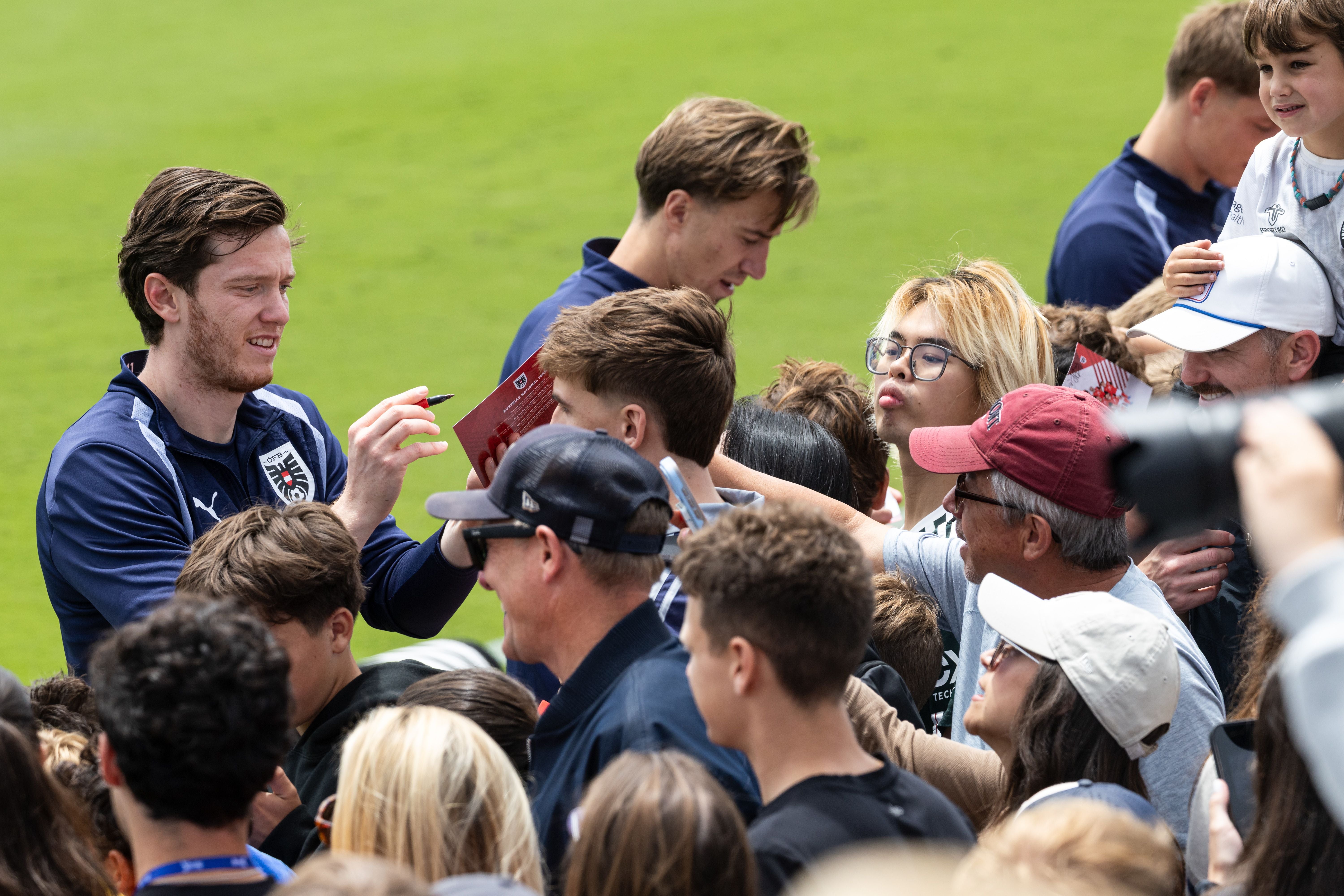 Michael Gregoritsch im dunkelblauen ÖFB-Trainingsanzug schreibt mit einem Stift ein Autogramm für Fans, die sich um ihn drängen.