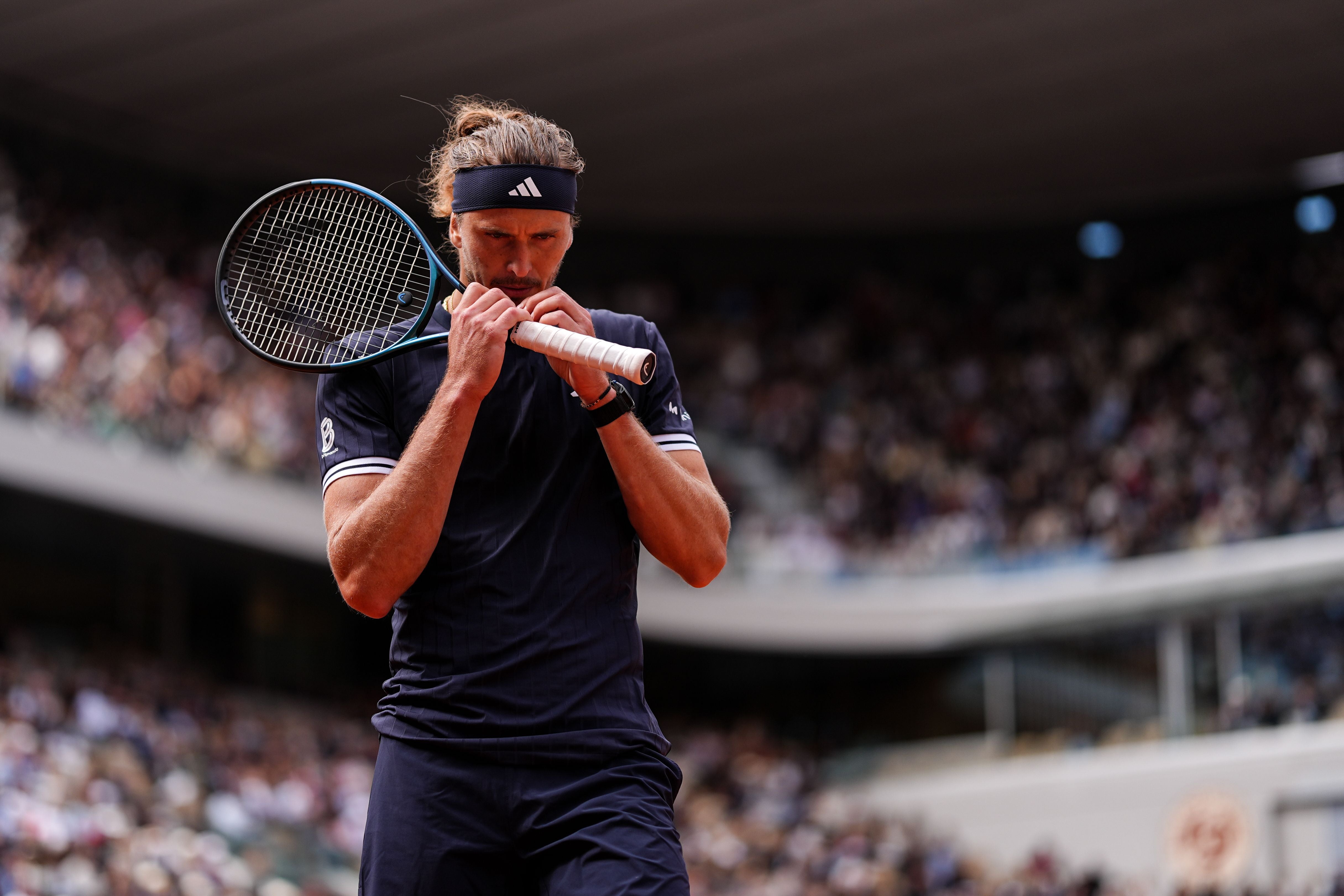 Der Tennisspieler Alexander Zverev steht in dunkelblauem Outfit und passendem Stirnband auf einem Sandplatz, blickt konzentriert nach unten und hält den Griff seines Tennisschlägers mit beiden Händen vor das Gesicht, während im Hintergrund die vollbesetzten Zuschauertribünen eines Stadions unscharf zu sehen sind.