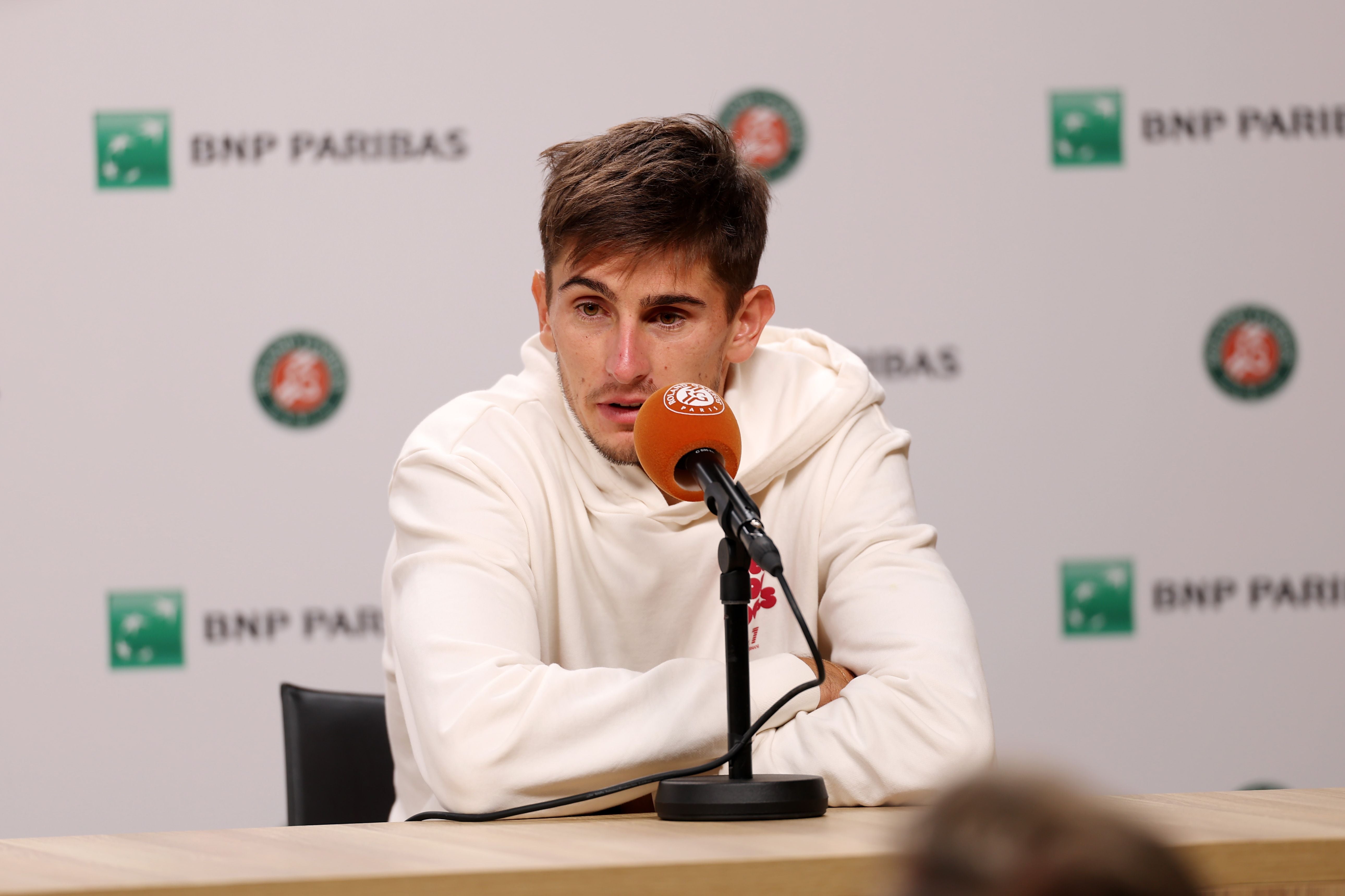 Der italienische Tennis-Profi Matteo Arnaldi sitzt in einem weißen Kapuzenpullover an einem Holztisch vor einer Pressewand mit Sponsorenlogos und spricht in ein orangefarbenes Mikrofon. Arnaldi musste sein Match in Paris wegen einer Viruserkrankung absagen und verpasste damit die Chance auf das Endspiel gegen Alexander Zverev.