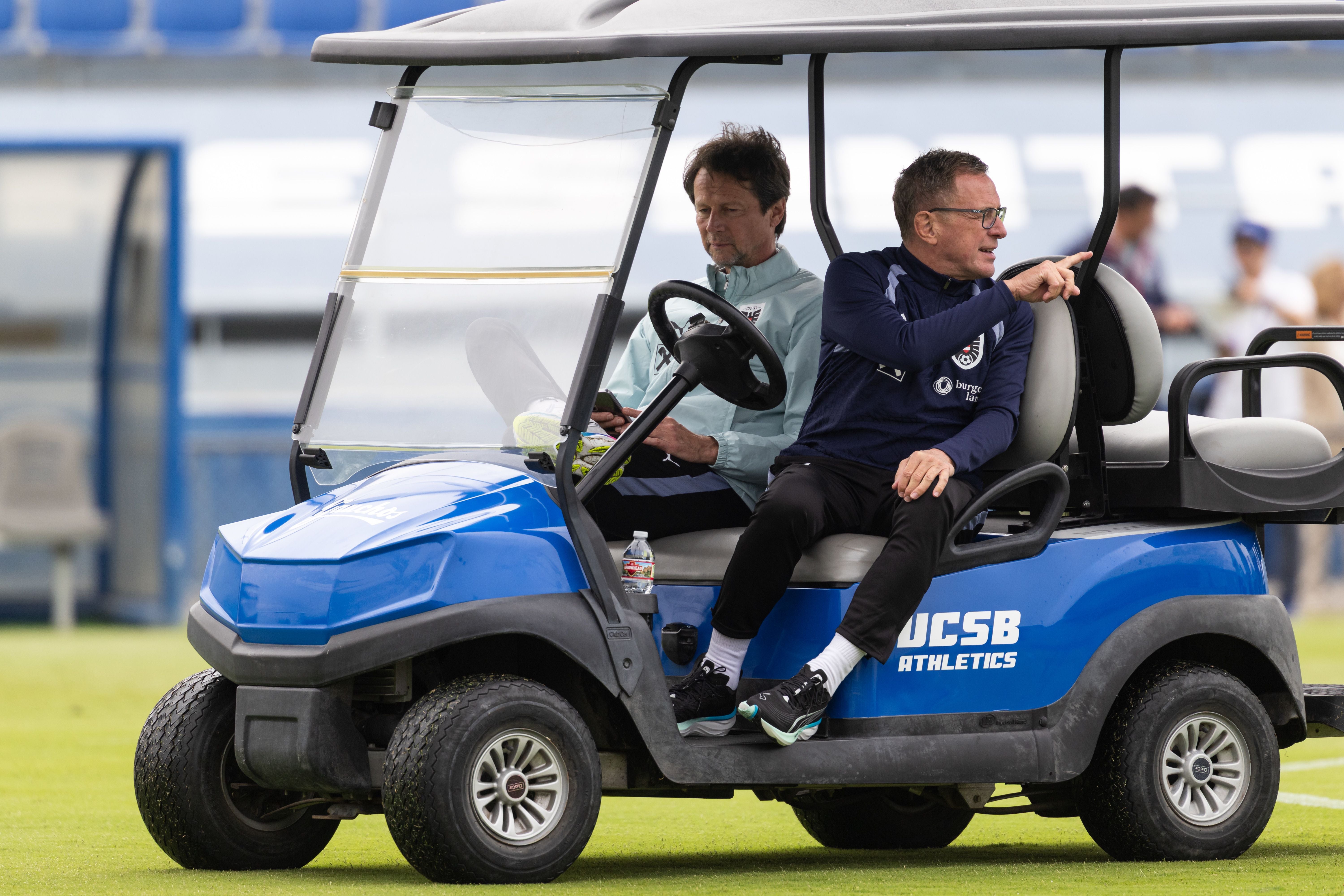 Teamchef Ralf Rangnick und Sportdirektor Peter Schöttel im ÖFB-Trainingsgewand sitzen nebeneinander in einem kleinen, blau-schwarzen Golfwagen auf einem grünen Fußballplatz, während der Teamchef mit Brille nach hinten deutet.