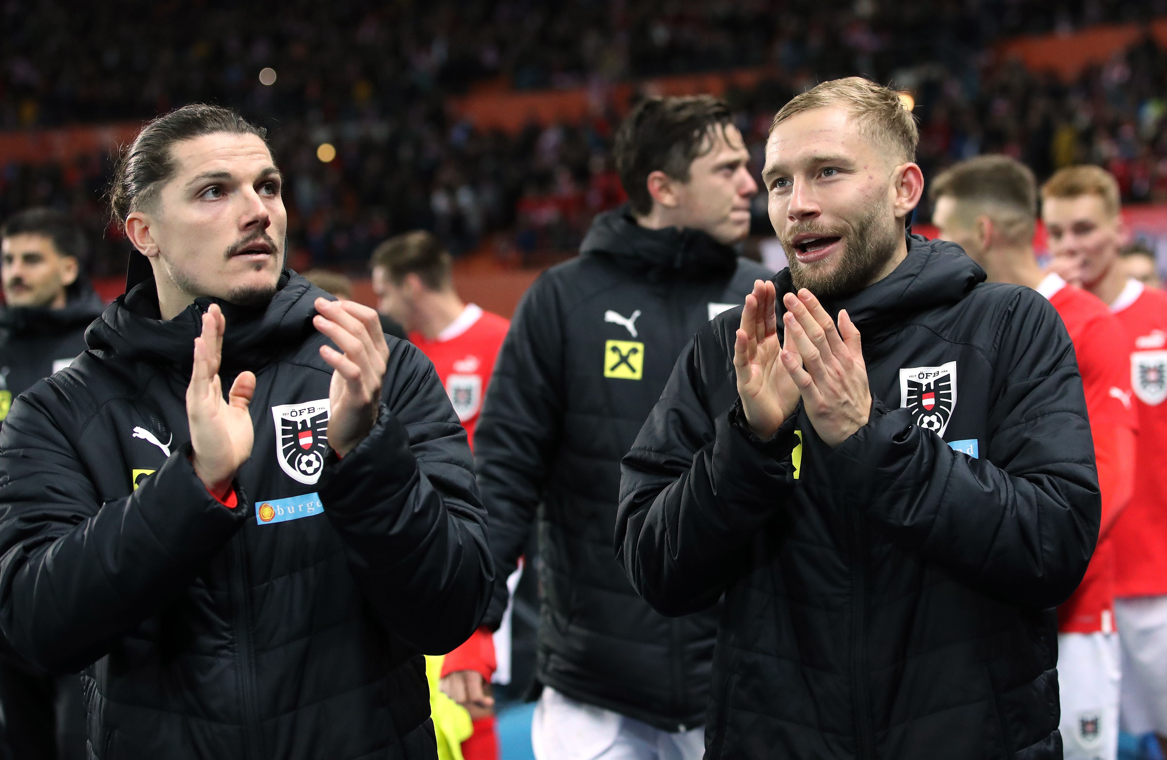 Marcel Sabitzer und Konrad Laimer stehen in schwarzen ÖFB-Jacken nebeneinander auf dem Rasen und klatschen in die Hände. Im Hintergrund sind weitere Spieler in roten ÖFB-Trikots und Zuschauerränge im Stadion zu sehen.