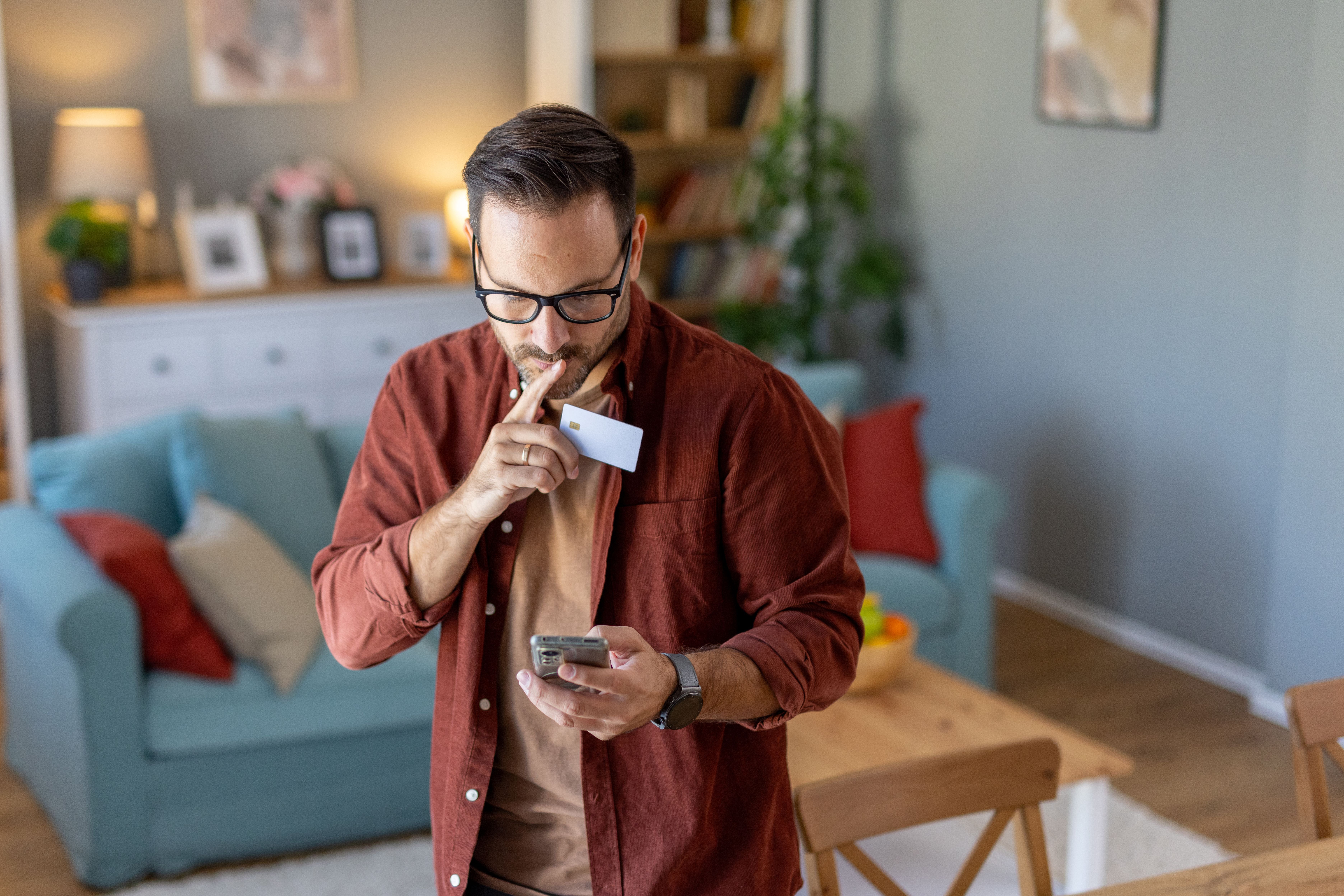 Thoughtfully, the man holds a credit card and phone, deep in consideration while surrounded by a stylish and engaging living atmosphere.