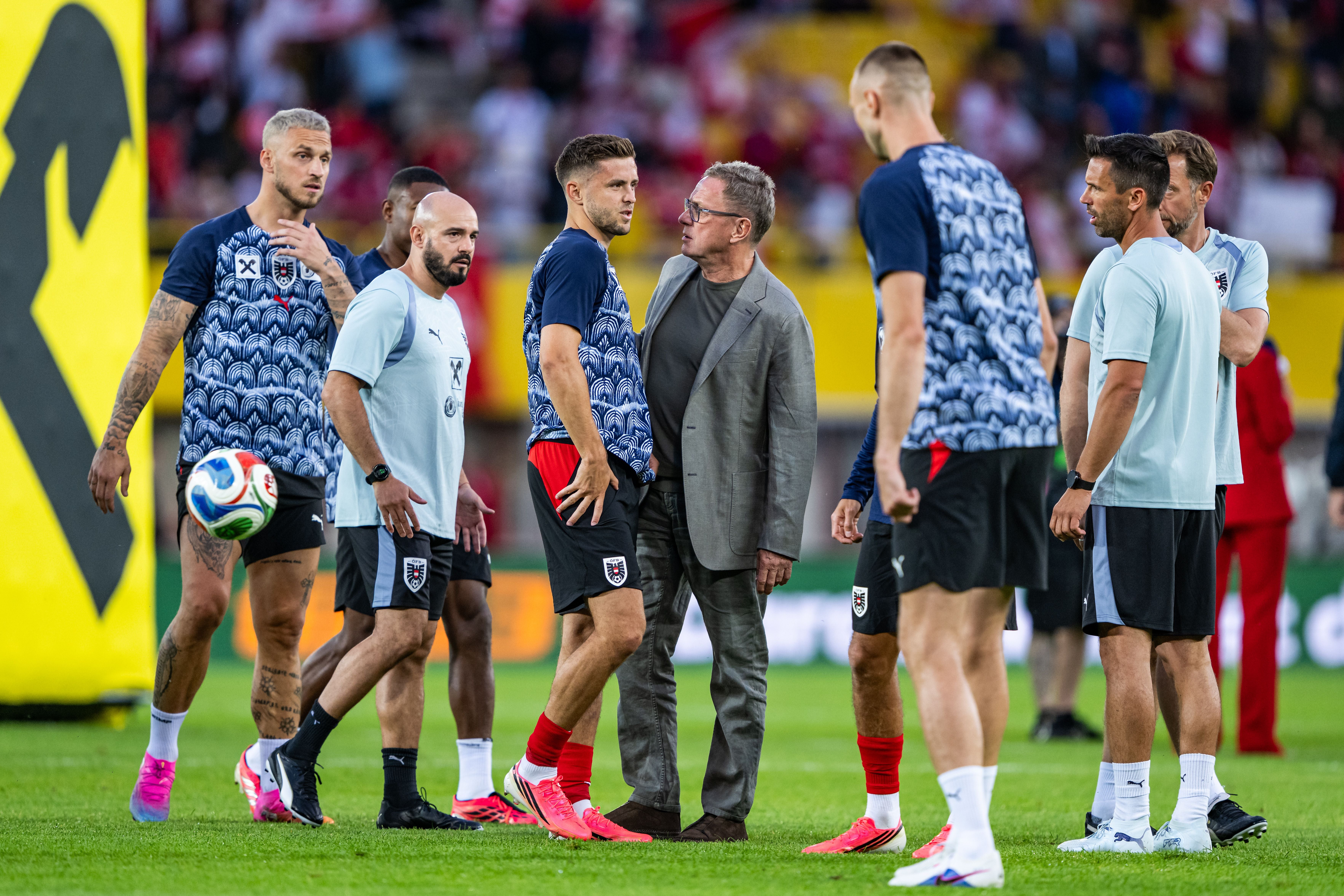 Mehrere Spieler und Betreuer der österreichischen Fußball-Nationalmannschaft stehen während einer Trainingseinheit oder Aufwärmphase auf dem Rasen eines Stadions. Im Zentrum Österreichs Nationaltrainer Ralf Rangnick im Gespräch mit Christoph Baumgartner, der sich an den Oberschenkel greift. Links daneben steht Marko Arnautovic mit einem Ball am Fuß, während weitere Teammitglieder in Trainingskleidung die Szene umgeben. Im Hintergrund sind gut gefüllte Zuschauertribünen zu erkennen.