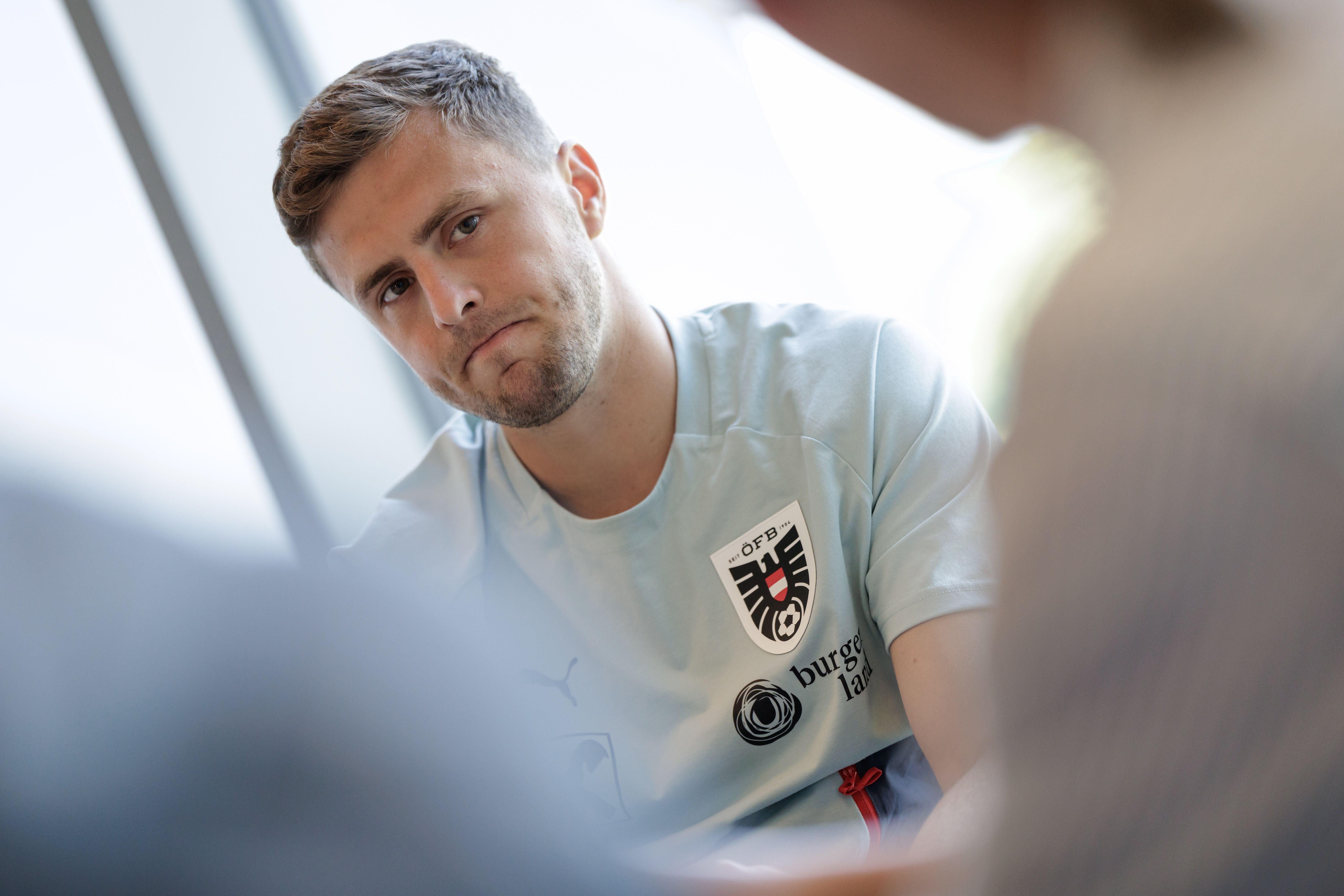 Christoph Baumgartner sitzt in einem hellen ÖFB-Trainingsshirt mit dem offiziellen Adler-Verbandswappen und dem Burgenland-Sponsorenlogo in einem Raum. Er blickt mit einem nachdenklichen, leicht traurigen Gesichtsausdruck direkt in die Kamera, während im Vordergrund unscharf die Silhouette einer anderen Person zu sehen ist.