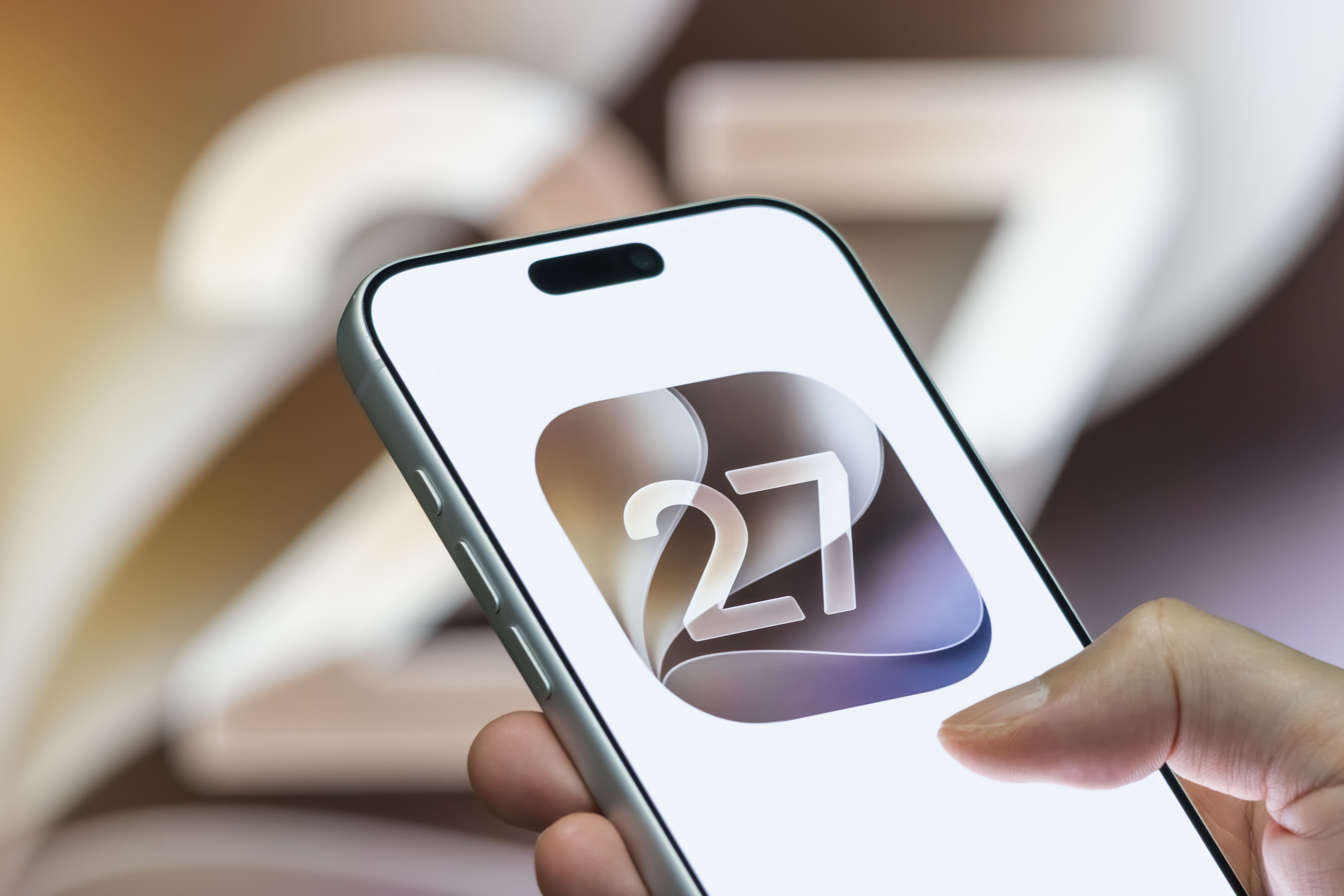 SHENZHEN, CHINA - JUNE 9: In this photo illustration, a person interacts with a smartphone displaying the Apple iOS 27 logo in front of a large iOS 27-themed background display on June 9, 2026 in Shenzhen, Guangdong Province, China. Apple unveiled iOS 27 during its Worldwide Developers Conference (WWDC) 2026, introducing updates focused on Apple Intelligence, enhancements to Siri, redesigned system apps and user interface improvements across the iPhone ecosystem. (Photo illustration by Cheng Xin/Getty Images)
