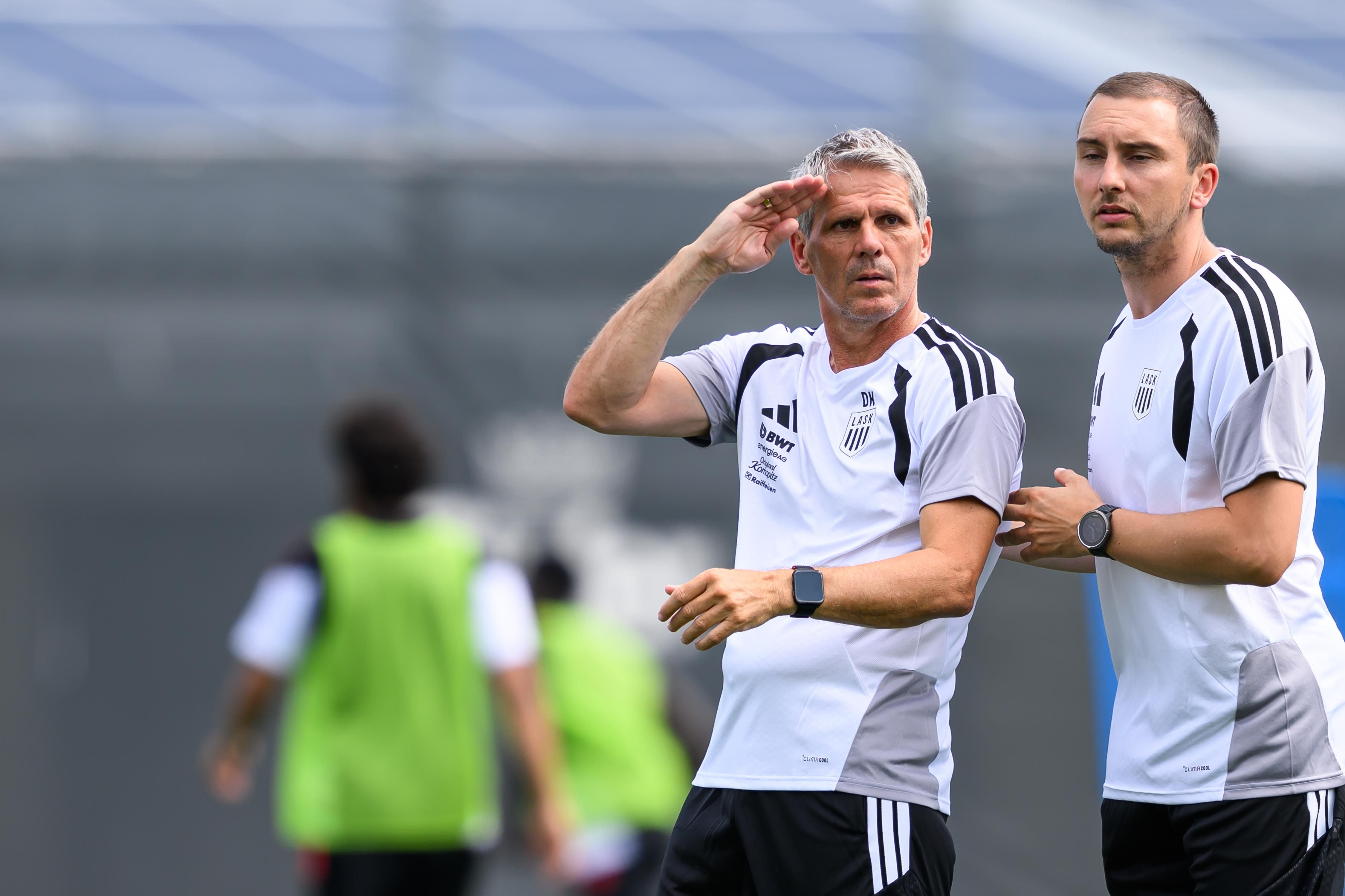LASK-Trainer Dietmar Kühbauer im weißen Trainingsshirt hält die Hand schützend an die Stirn und blickt konzentriert auf den Platz, während neben ihm ein Co-Trainer mit einer Sportuhr am Handgelenk steht.