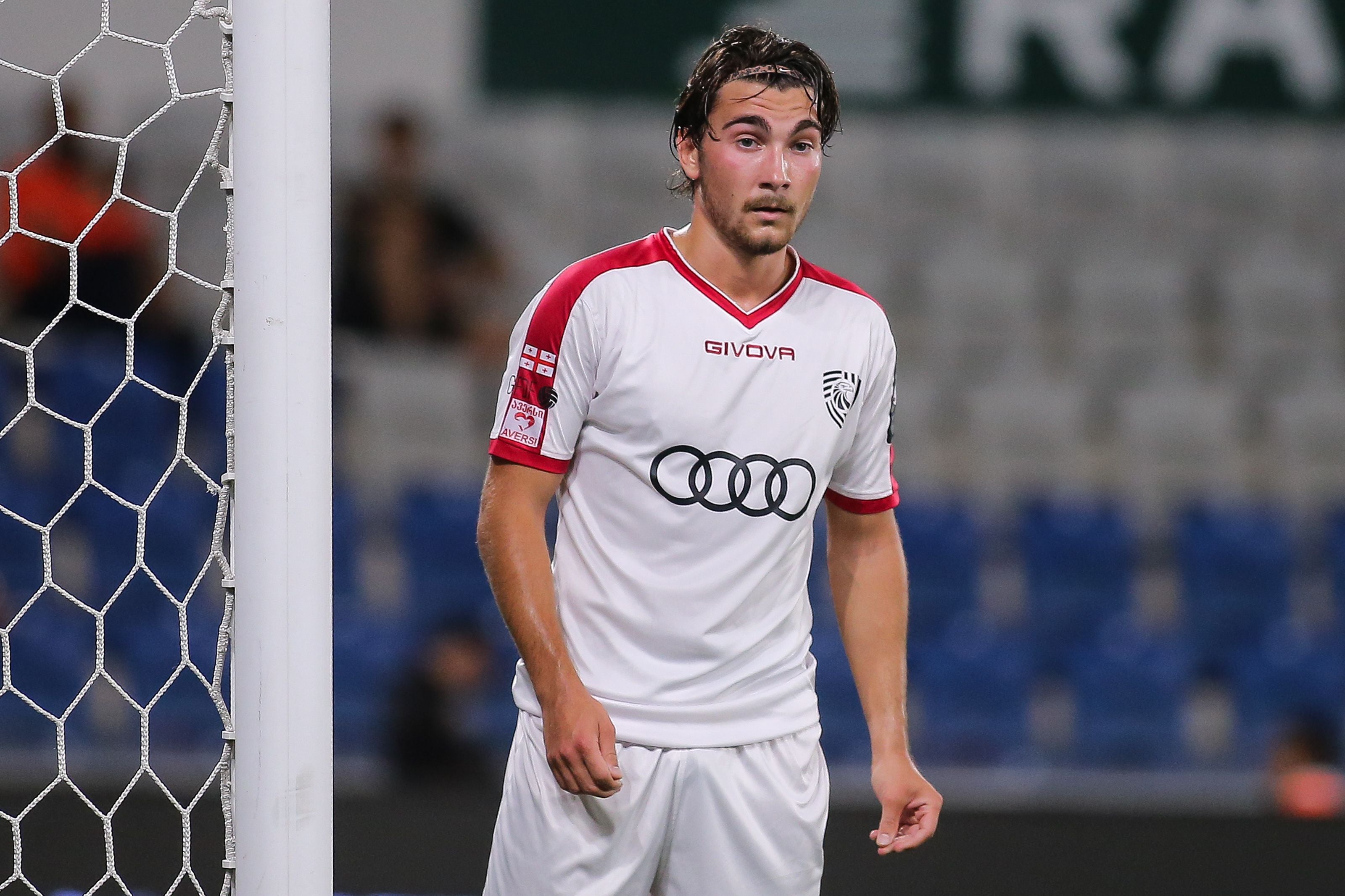 Der georgische Fußballspieler Otar Mamageishvili steht in einem weißen Trikot mit roten Ärmeln und einem Audi-Sponsorlogo auf der Brust nachdenklich auf dem Spielfeld neben dem Tornetz.