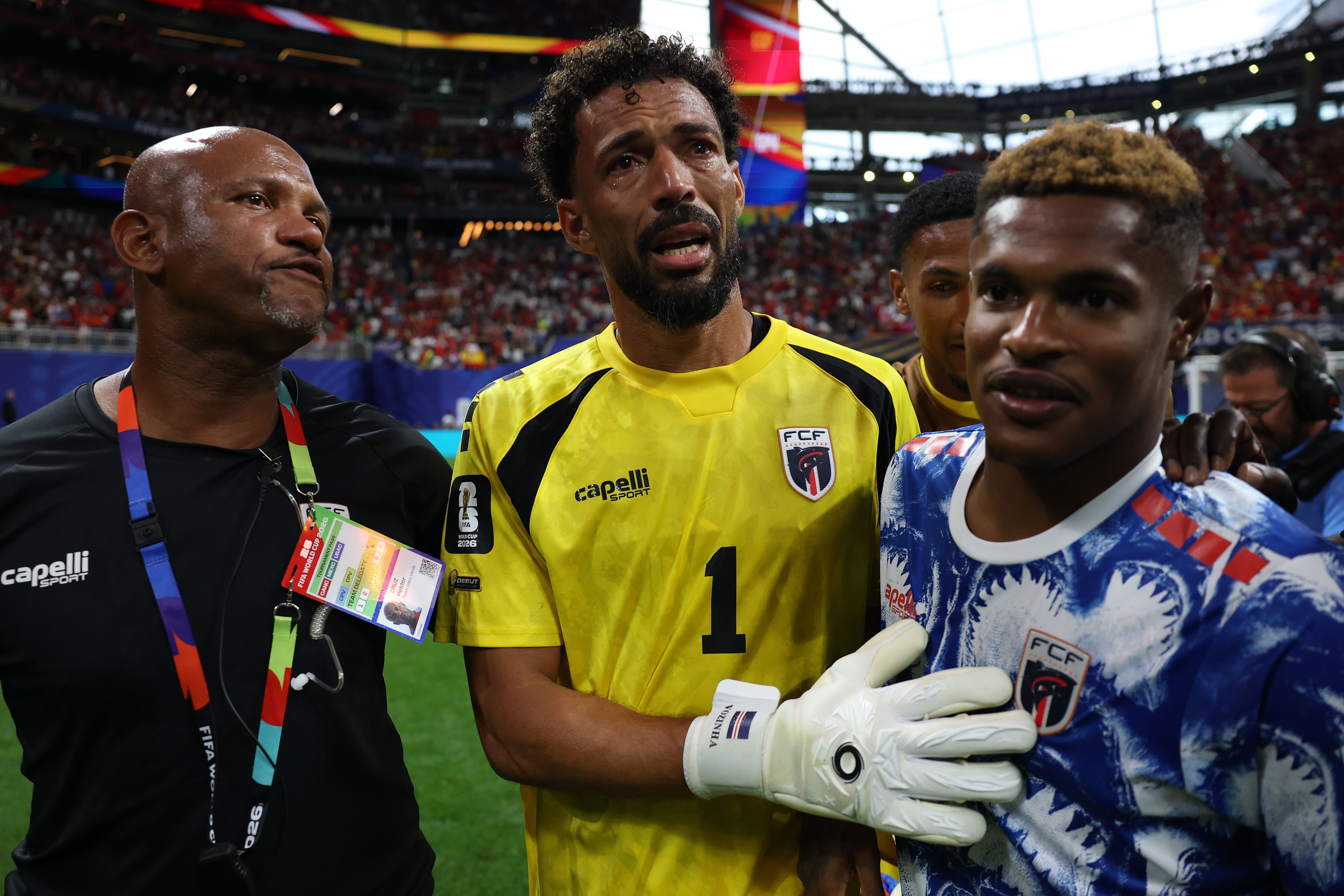 Kap-Verde-Torhüter Vozinha weint in einem gelben Trikot mit der Nummer eins auf dem Fußballplatz, während ihm ein Betreuer mit Akkreditierung und ein Teamkollege im blau-weißen Trikot zur Seite stehen.
