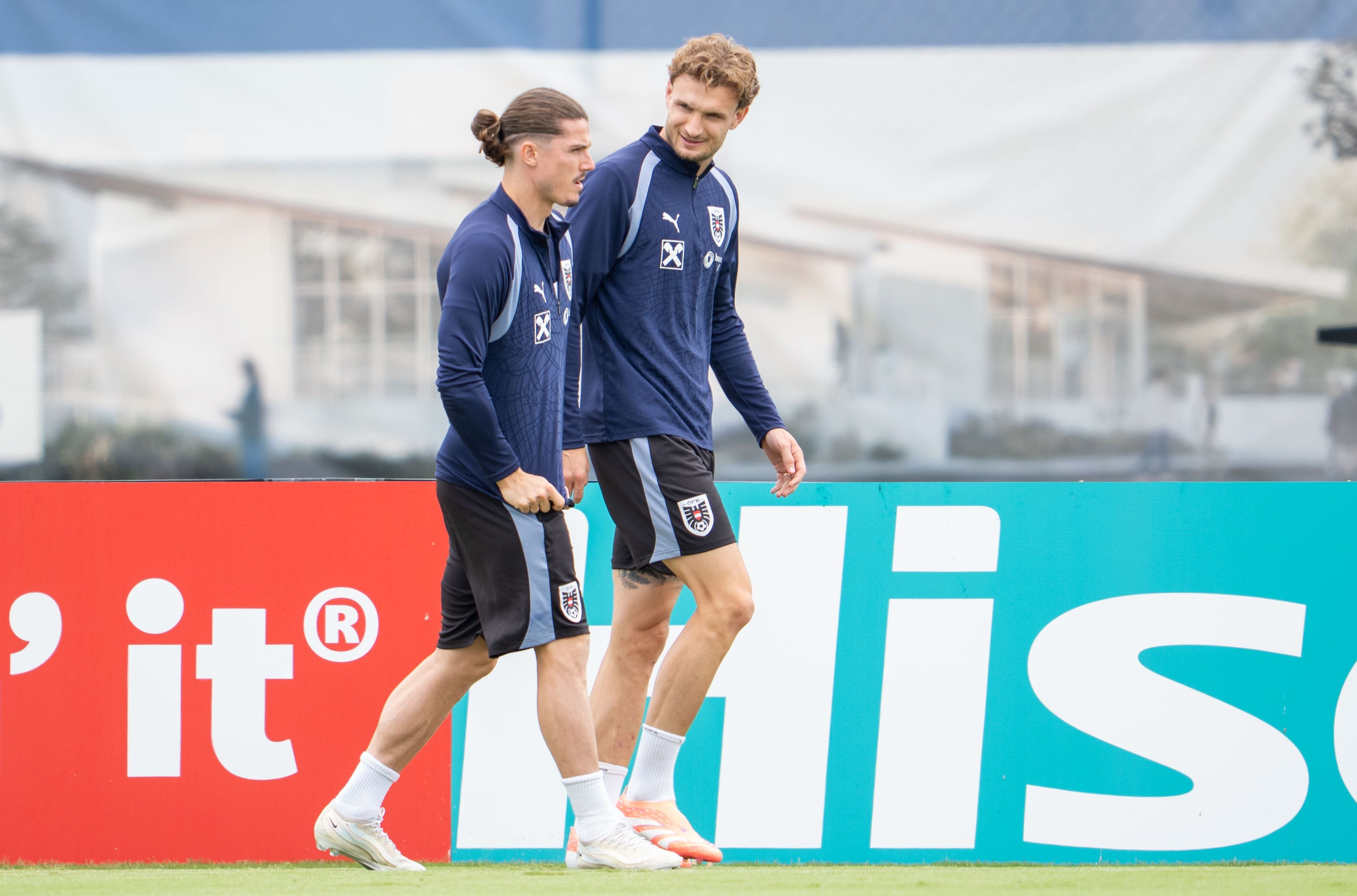 Zwei Fußballspieler im Trainingsanzug gehen nebeneinander auf einem Sportplatz entlang.