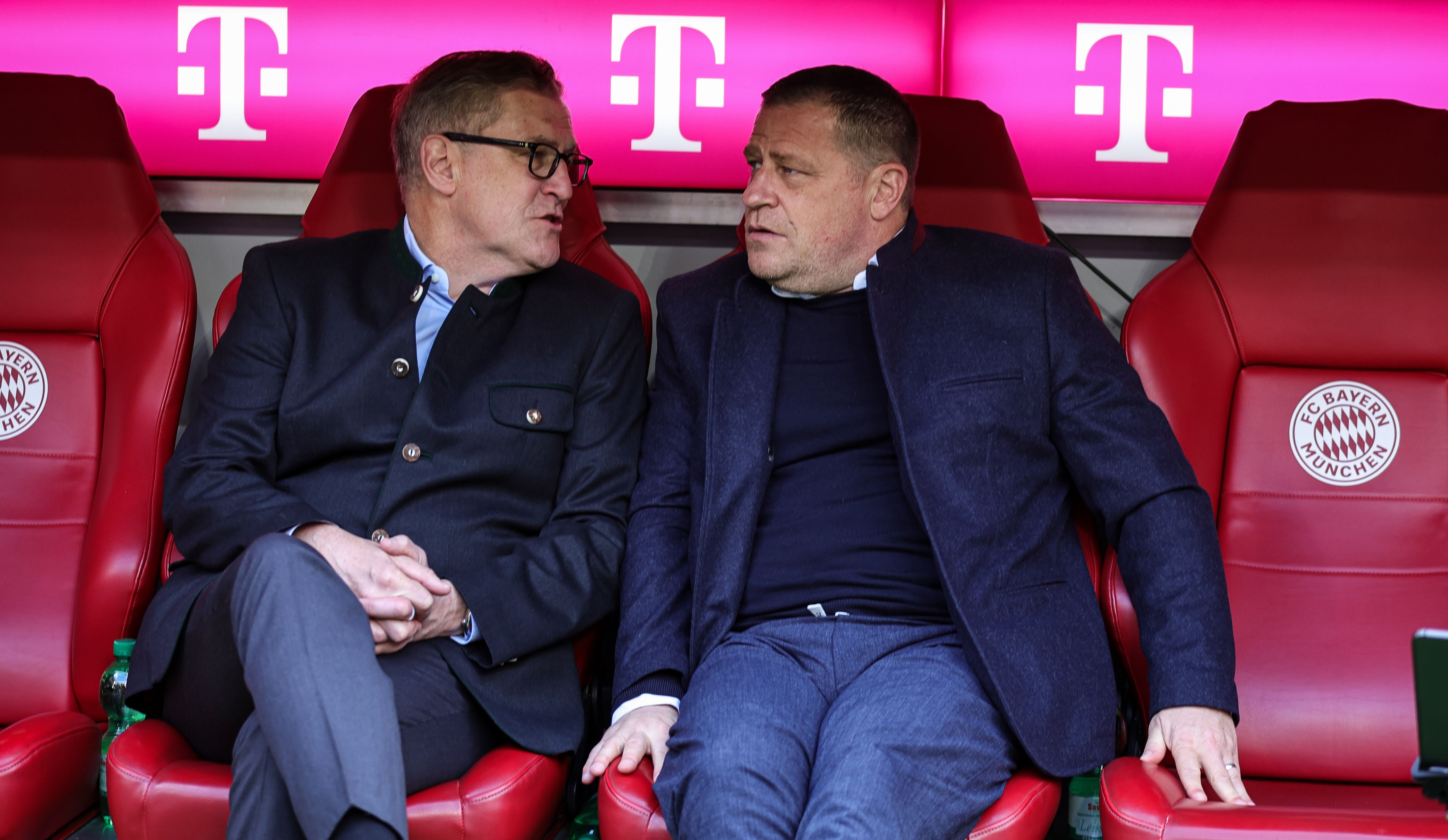 Zwei Männer im Anzug sitzen auf roten FC Bayern München-Sesseln in einem Stadion.