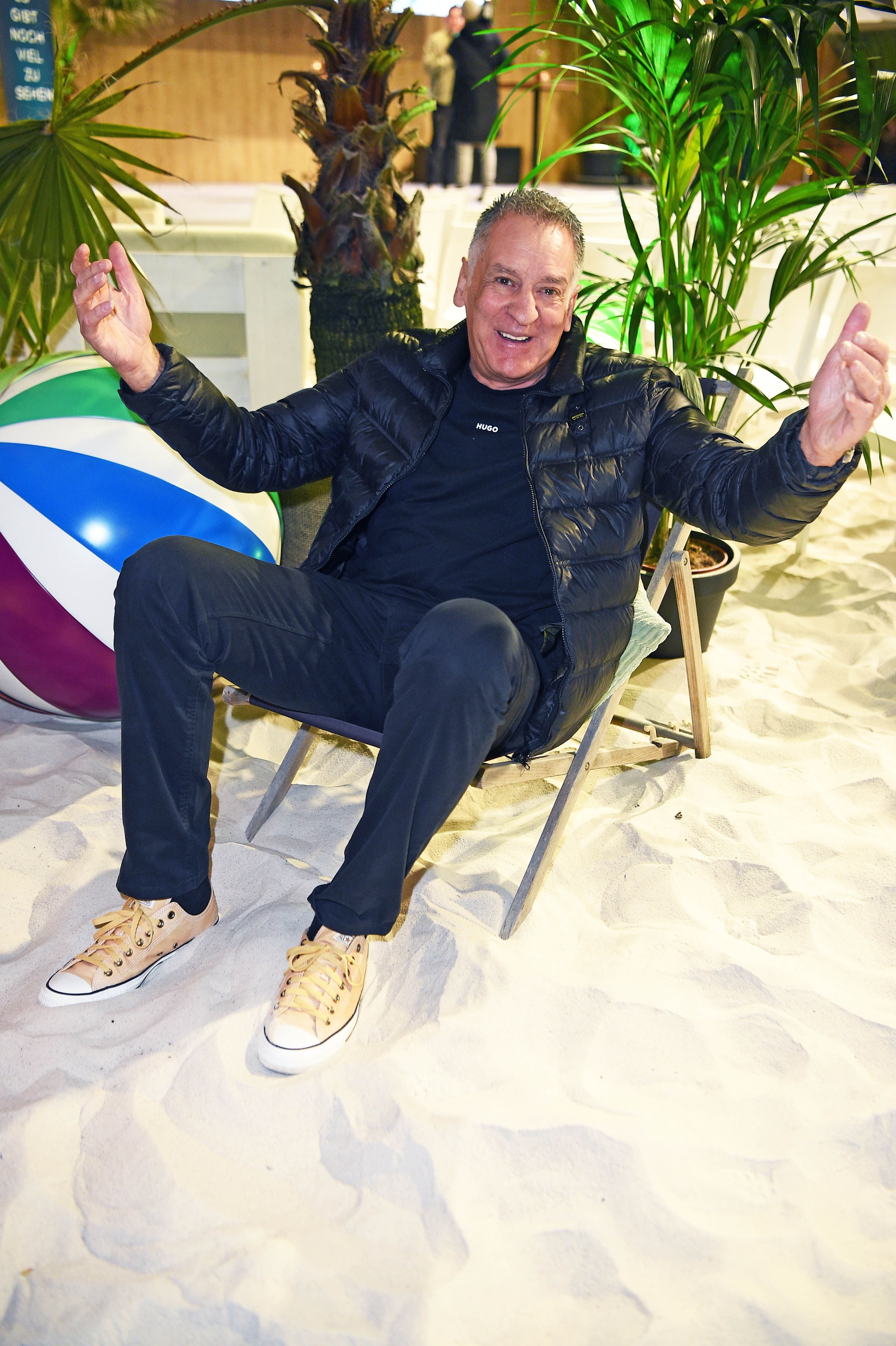 Ein Mann sitzt entspannt im Liegestuhl am Sandstrand, umgeben von Palmen und einem Wasserball.