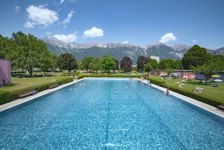 Großes Freibad mit Menschen, Liegewiese und Bergen im Hintergrund unter blauem Himmel.