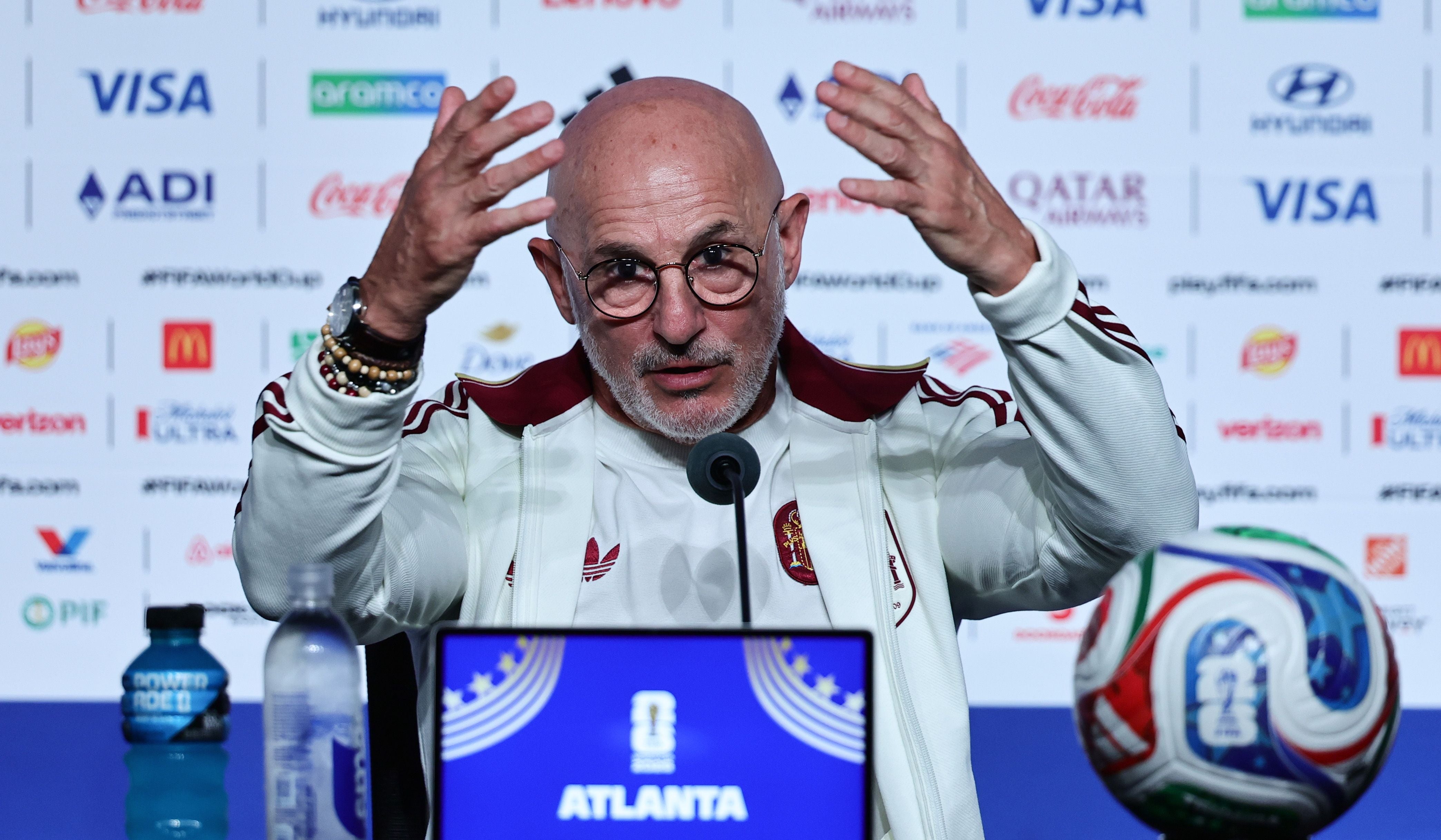 Ein Mann sitzt auf einer Pressekonferenz mit Mikrofon, Wasserflaschen und Fußball vor sich.
