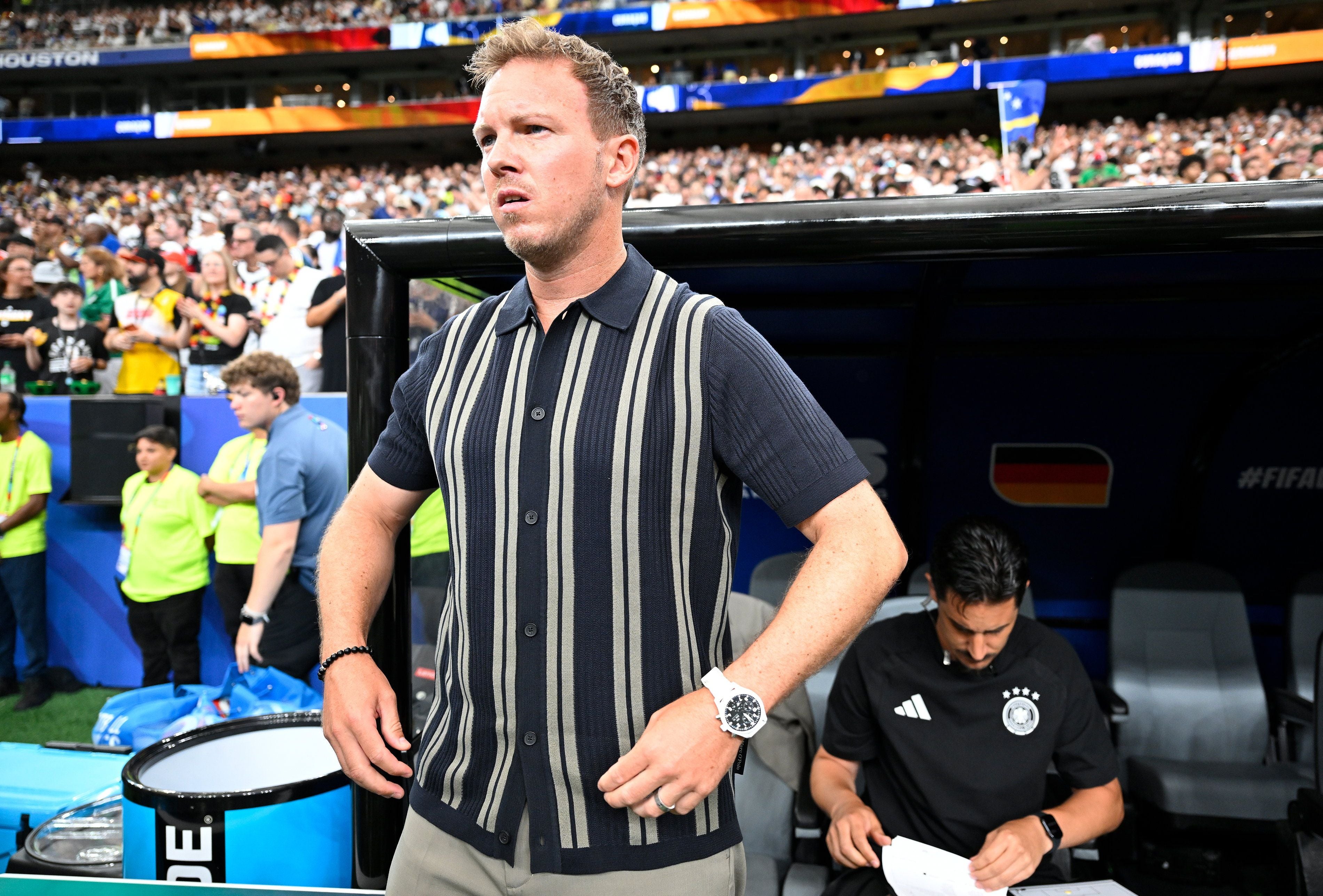 Ein Fußballtrainer steht vor der Bank während eines Spiels, im Hintergrund sieht man Zuschauer.