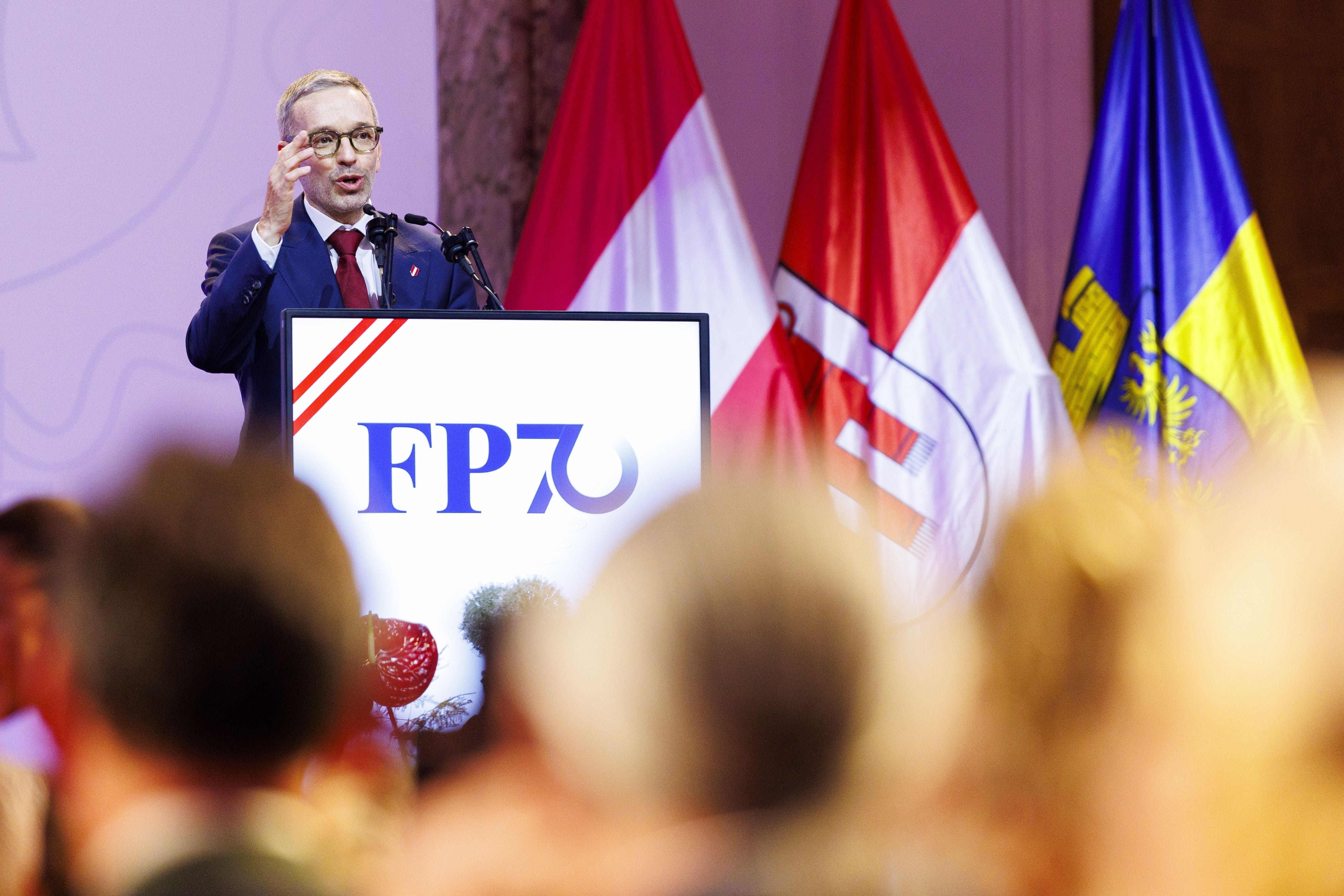 Ein Mann spricht bei einer Veranstaltung der FPÖ vor Fahnen und Publikum am Rednerpult.