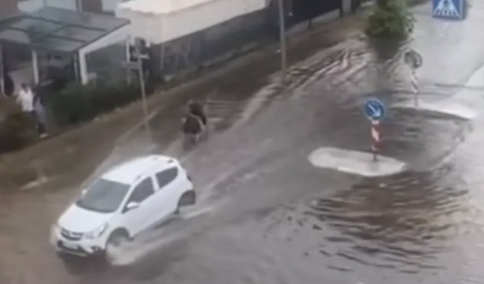 Ein weißes Auto fährt durch eine überflutete Straße in Berlin, zwei Menschen gehen daneben.