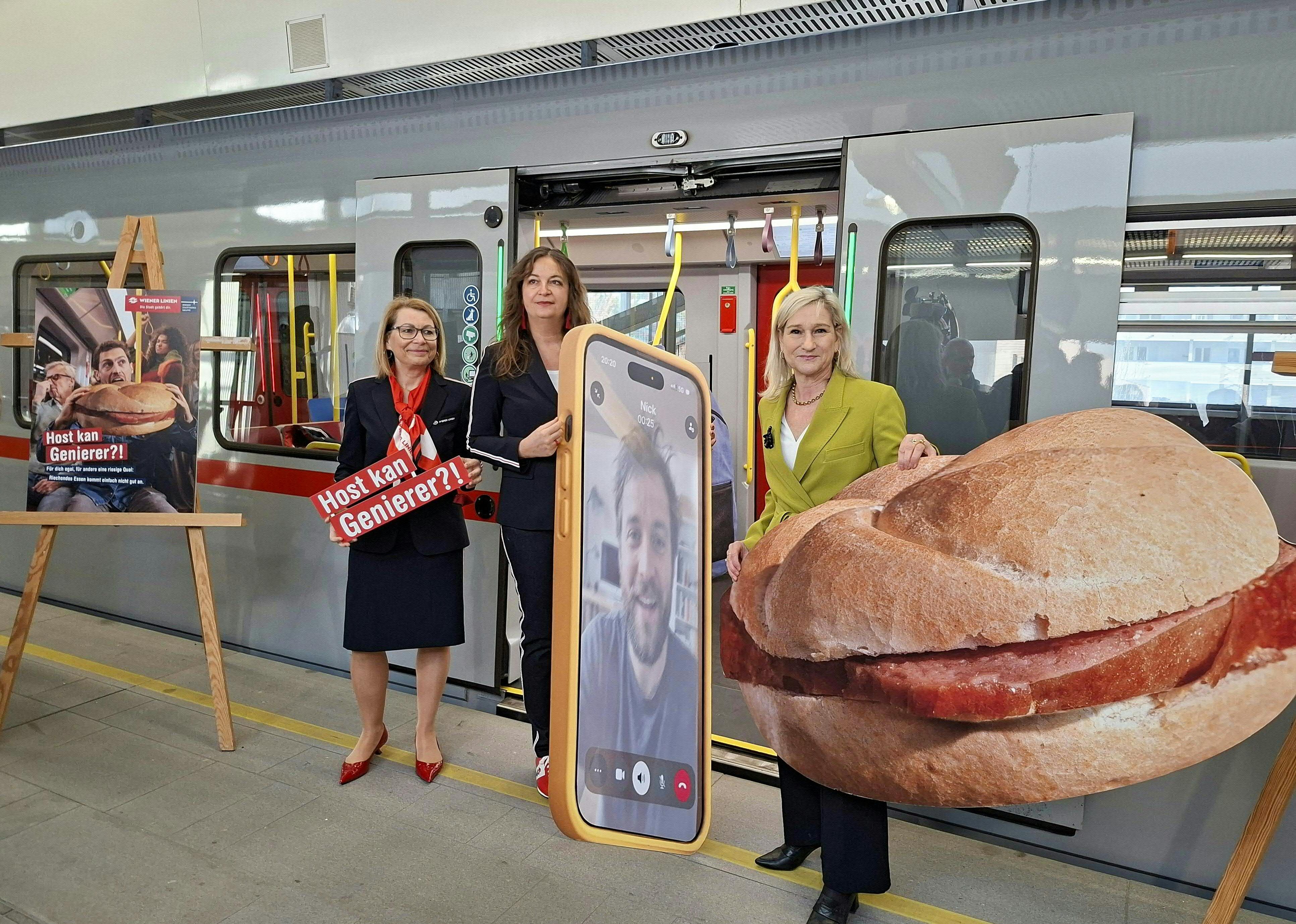 Drei Frauen stehen vor einer U-Bahn mit großen Pappaufstellern, darunter ein Handy und ein Wurstsemmel.
