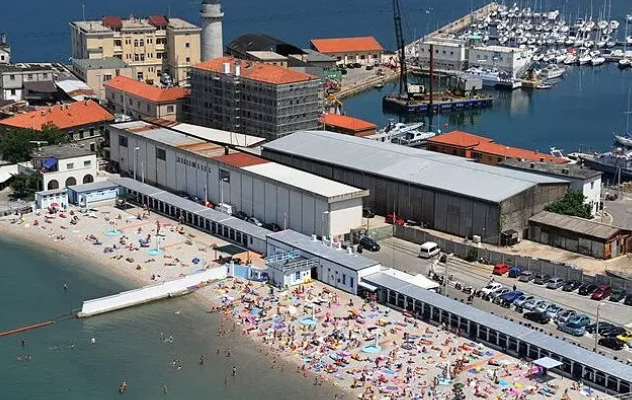Luftaufnahme vom Strandbad El Pedocin mit vielen Badegästen und Gebäuden am Hafen.