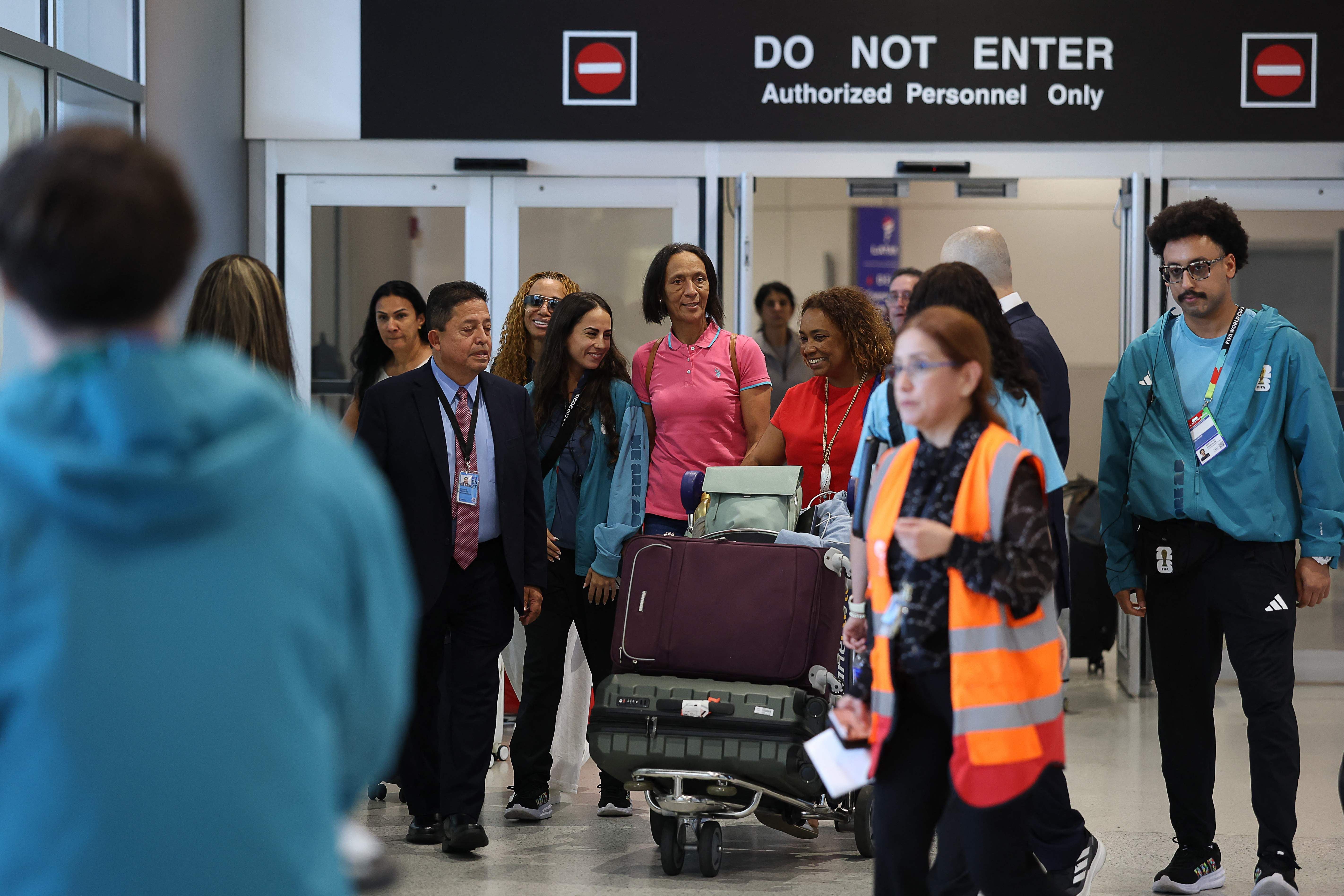 Mehrere Personen mit Gepäck verlassen einen Flughafen-Terminal, im Hintergrund ein „Do Not Enter“-Schild.