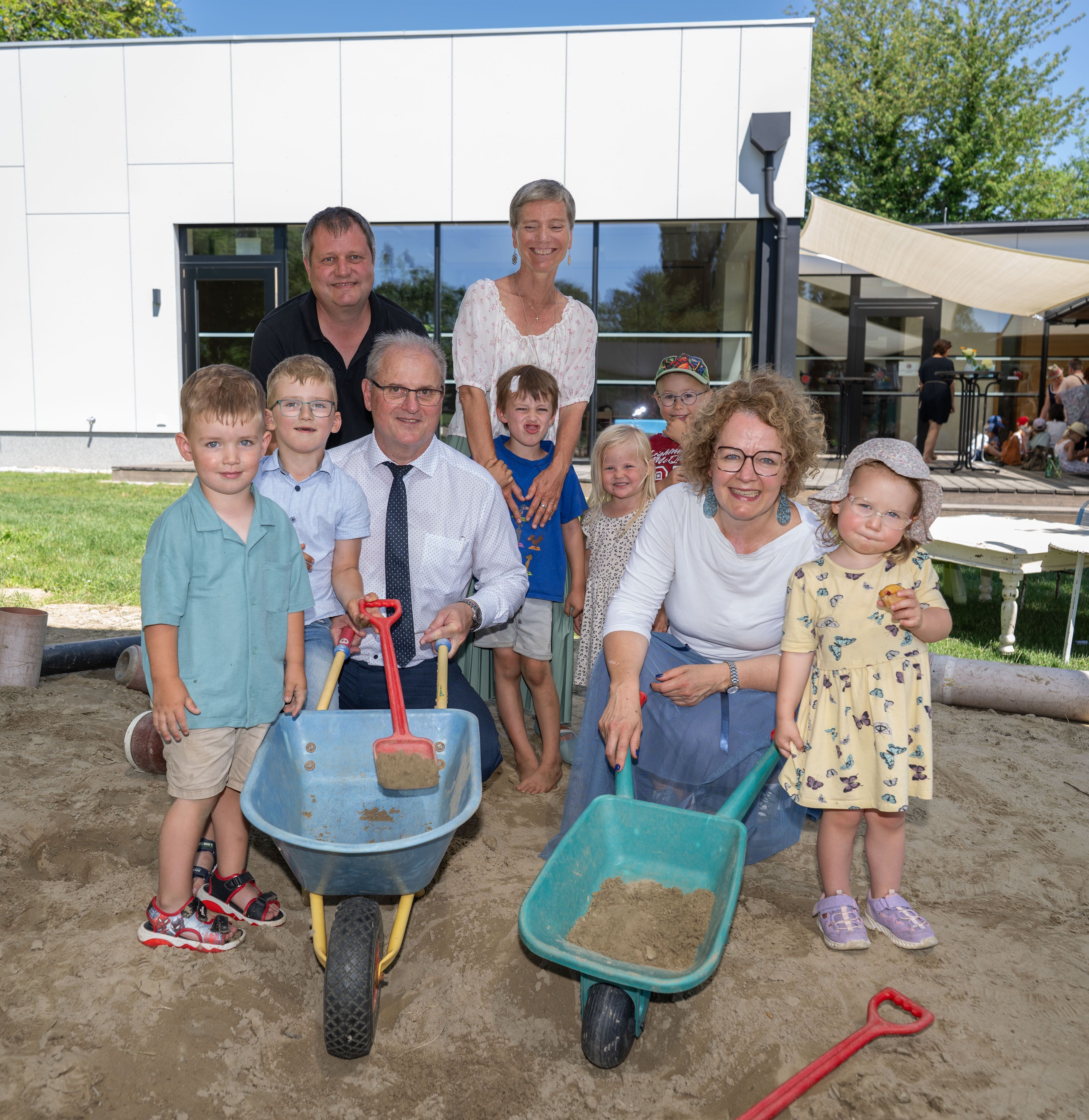 Gruppe von Erwachsenen und Kindern mit Schubkarren im Sandkasten vor einem modernen Gebäude.