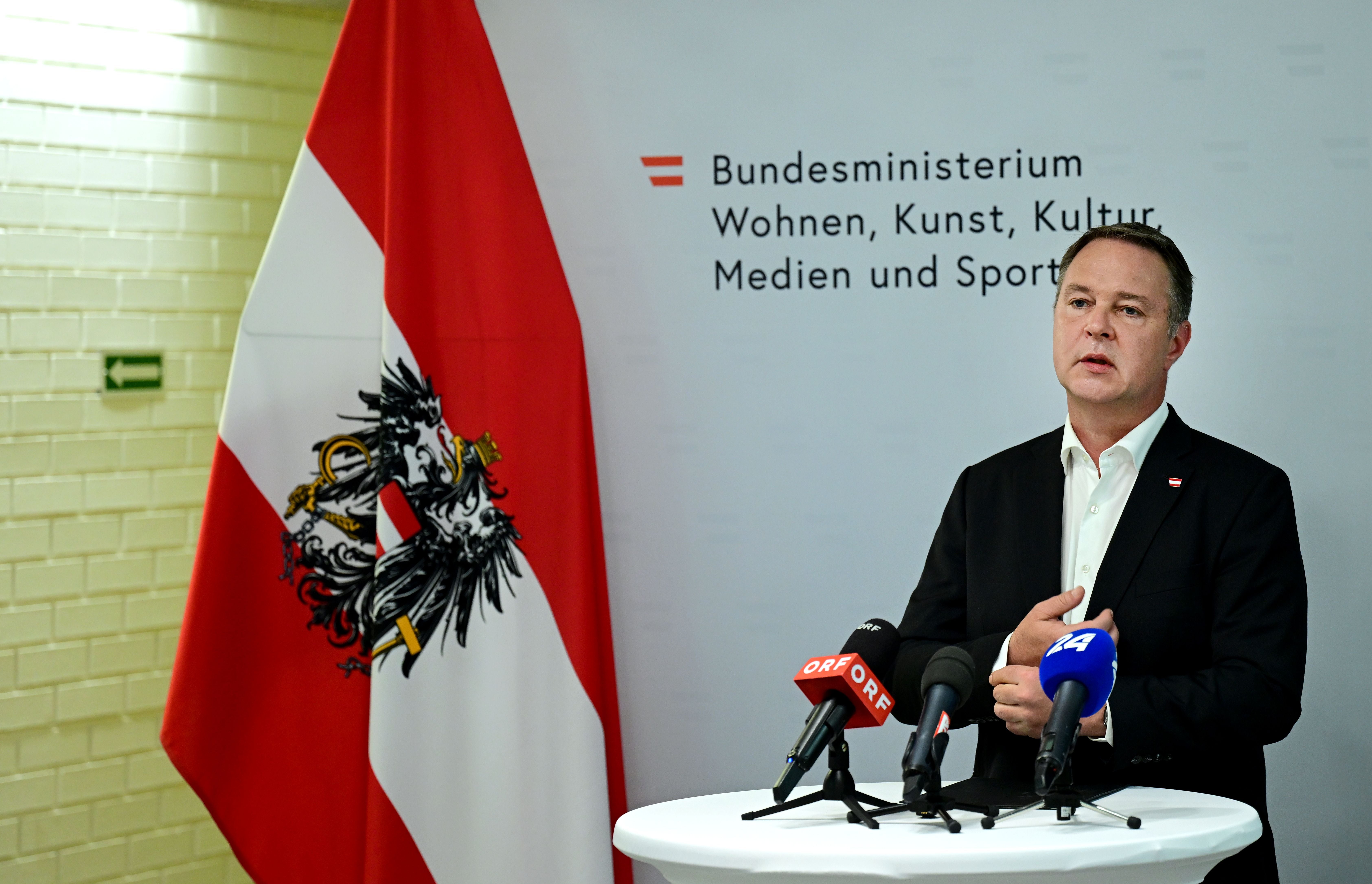 Ein Mann spricht bei einer Pressekonferenz vor einer Österreich-Fahne und mehreren Mikrofonen.