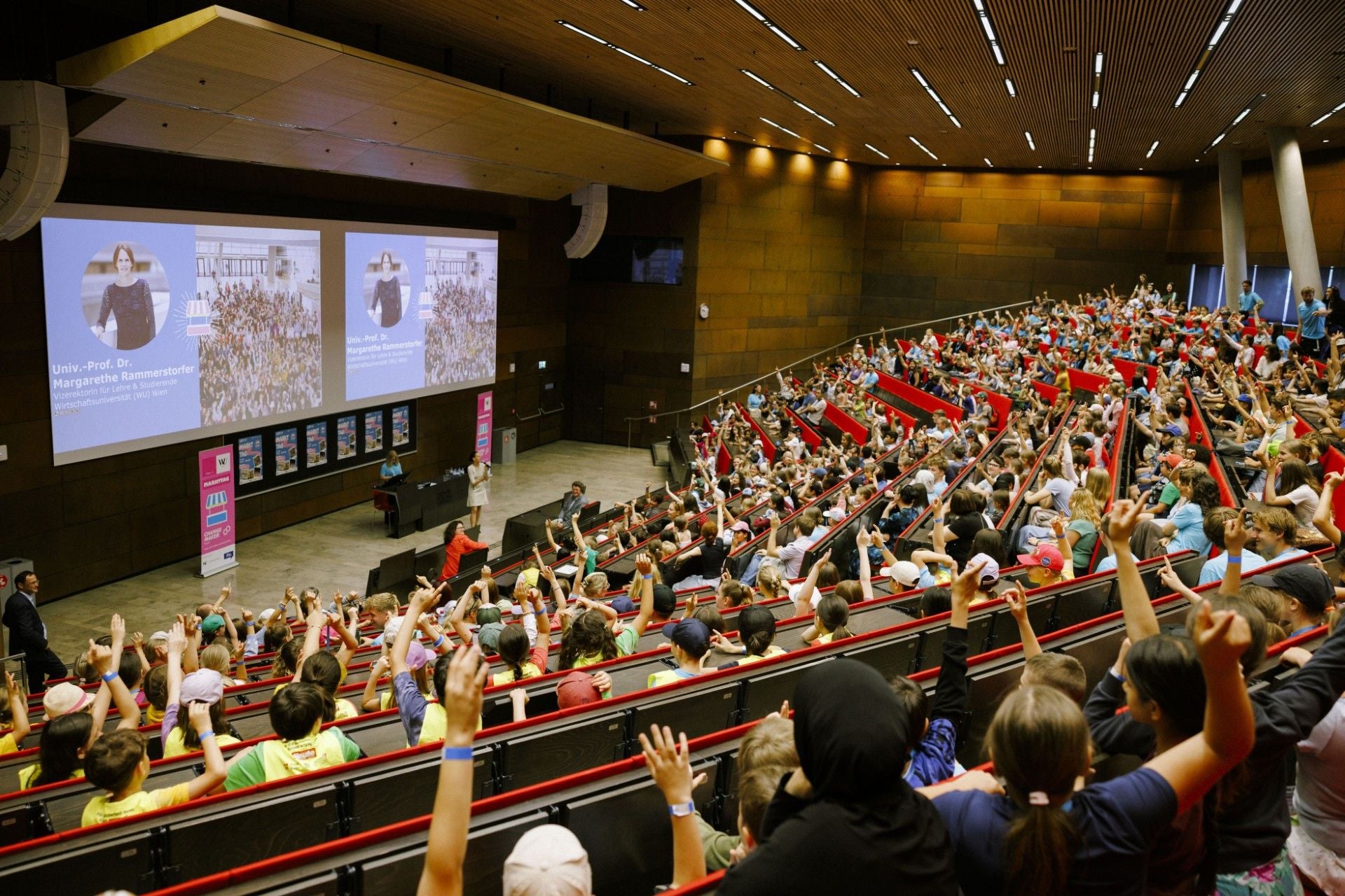 Viele Kinder sitzen in einem großen Hörsaal und heben die Hand bei einer Vorlesung im Audimax der WU.
