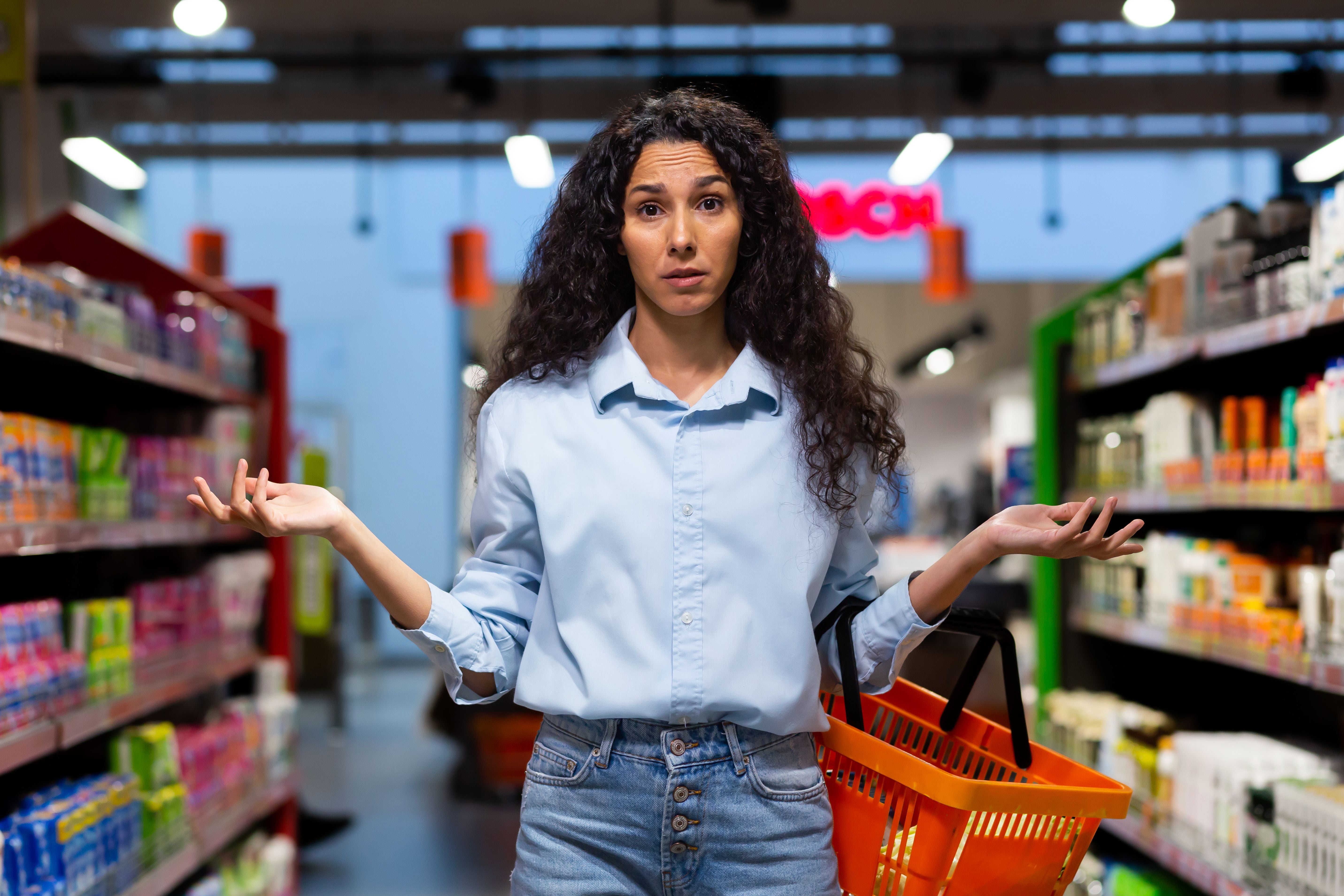 Junge Frau mit Einkaufskorb steht ratlos im Supermarkt zwischen Regalen mit Waren.