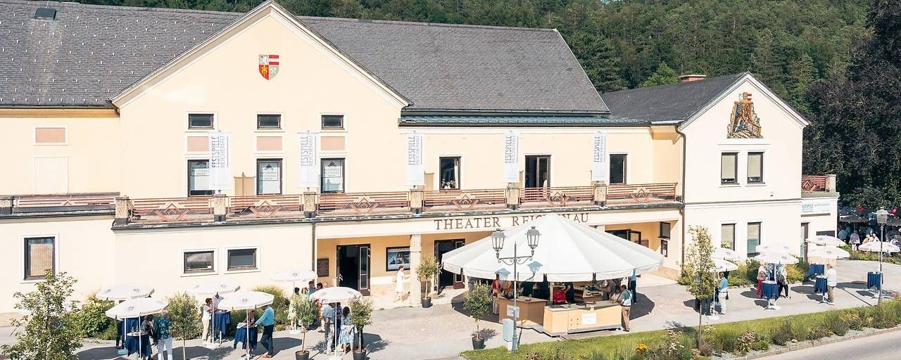 Das Theater Reichenau mit Besuchern und Sonnenschirmen vor dem Gebäude.