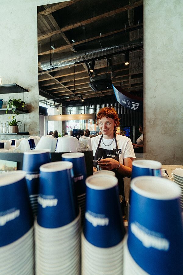Barista steht hinter einem Tresen mit gestapelten blauen Lavazza-Pappbechern im Café.