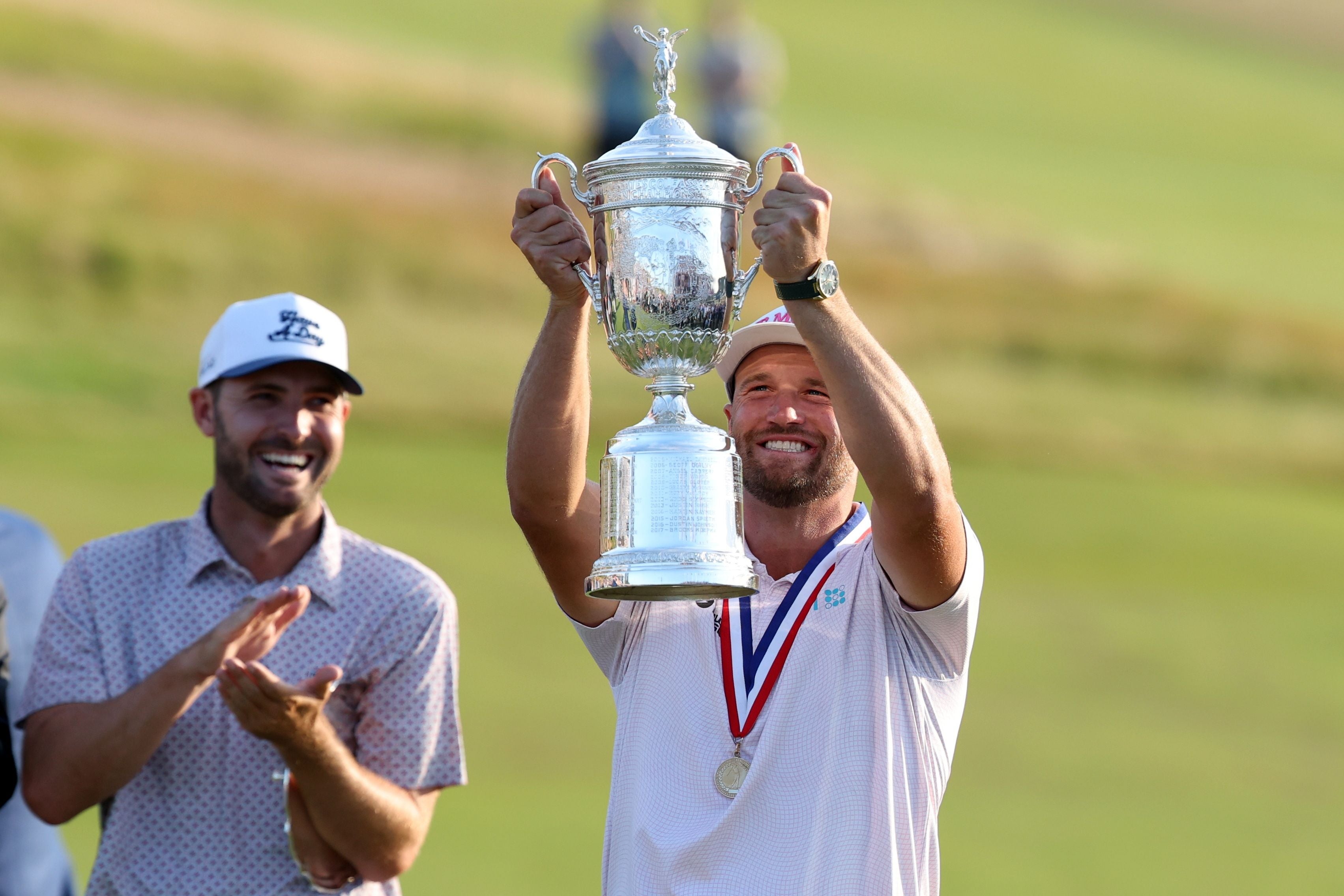 Ein Golfspieler hält einen großen Pokal hoch, daneben applaudiert sein Caddie auf dem Golfplatz.