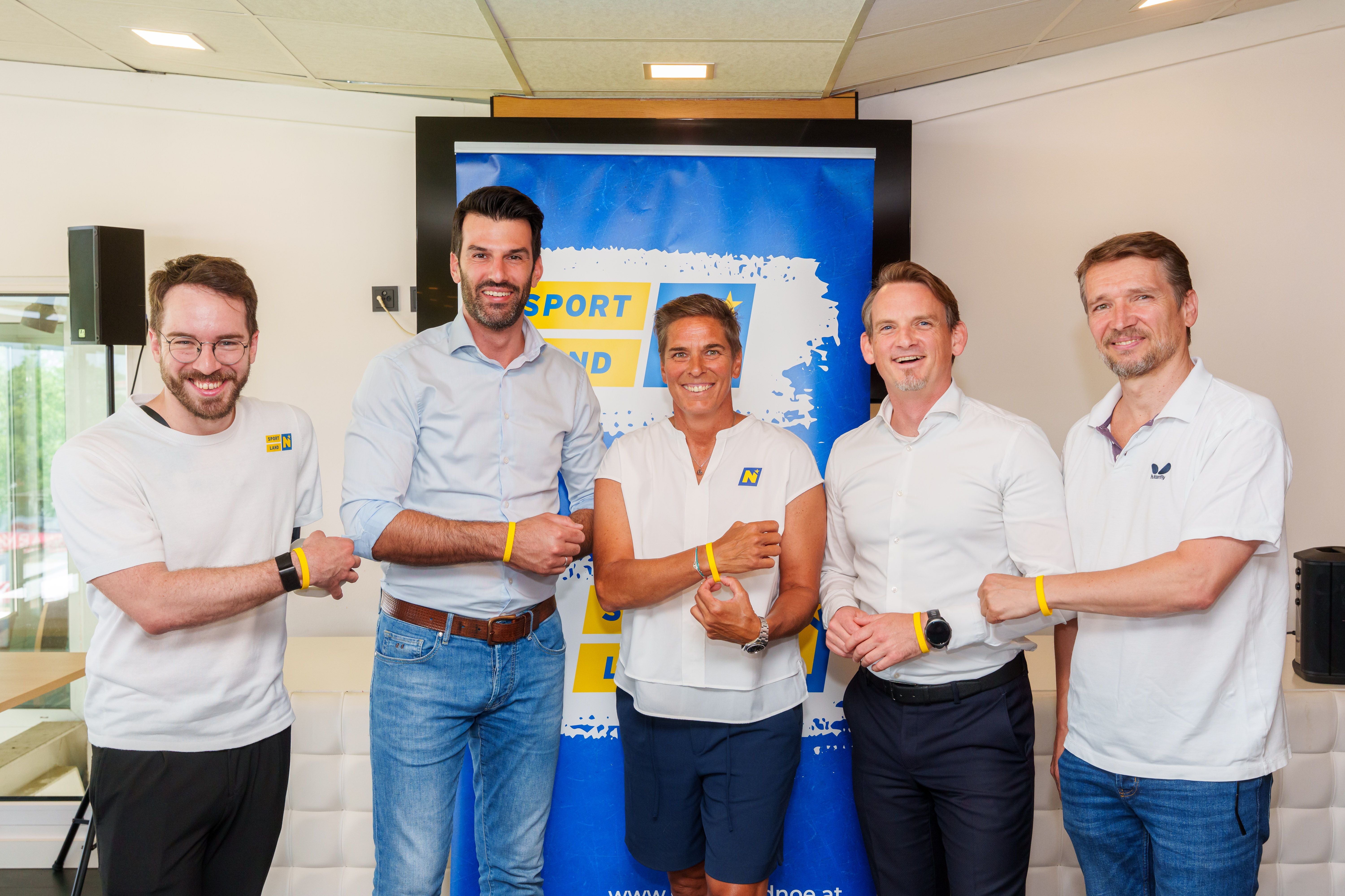 Fünf Personen posieren mit gelben Armbändern vor einem blauen Sportverband-Banner.