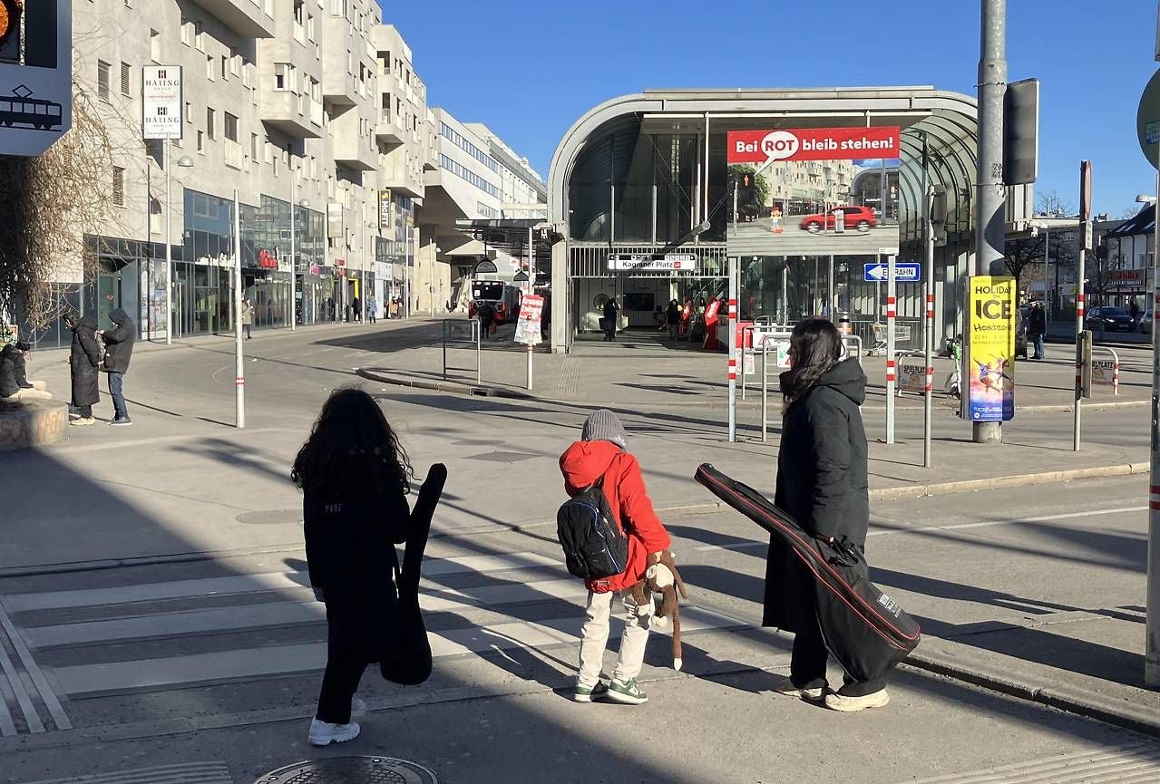 Drei Personen stehen an einem Zebrastreifen vor einer roten Fußgängerampel bei Sonnenschein.