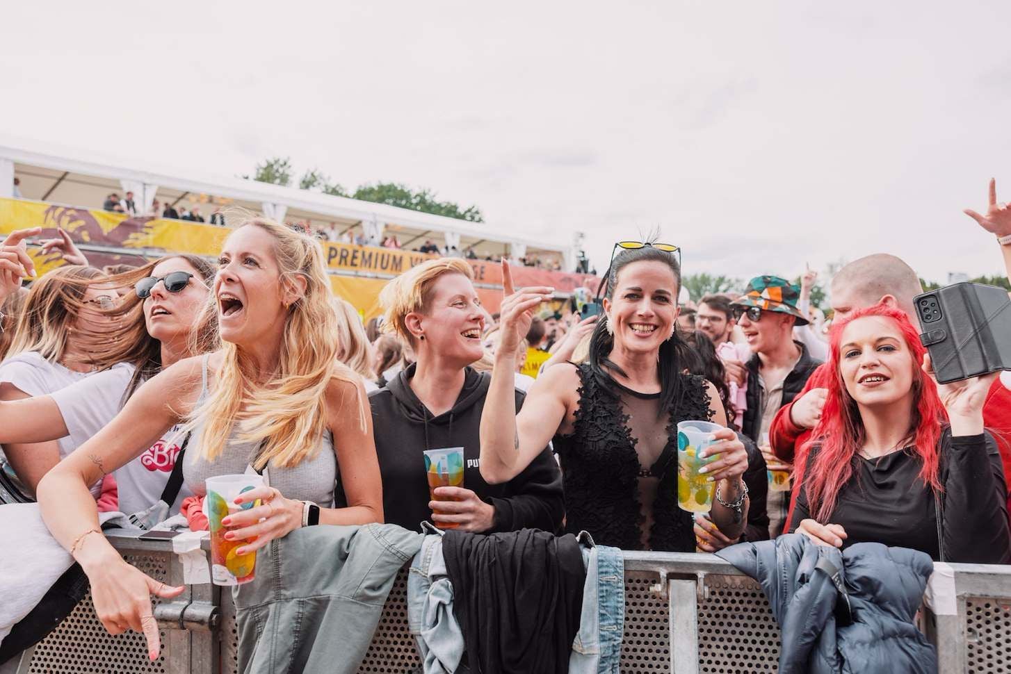 Mehrere Menschen feiern mit Getränken vor einer Absperrung auf einem Open-Air-Festival.