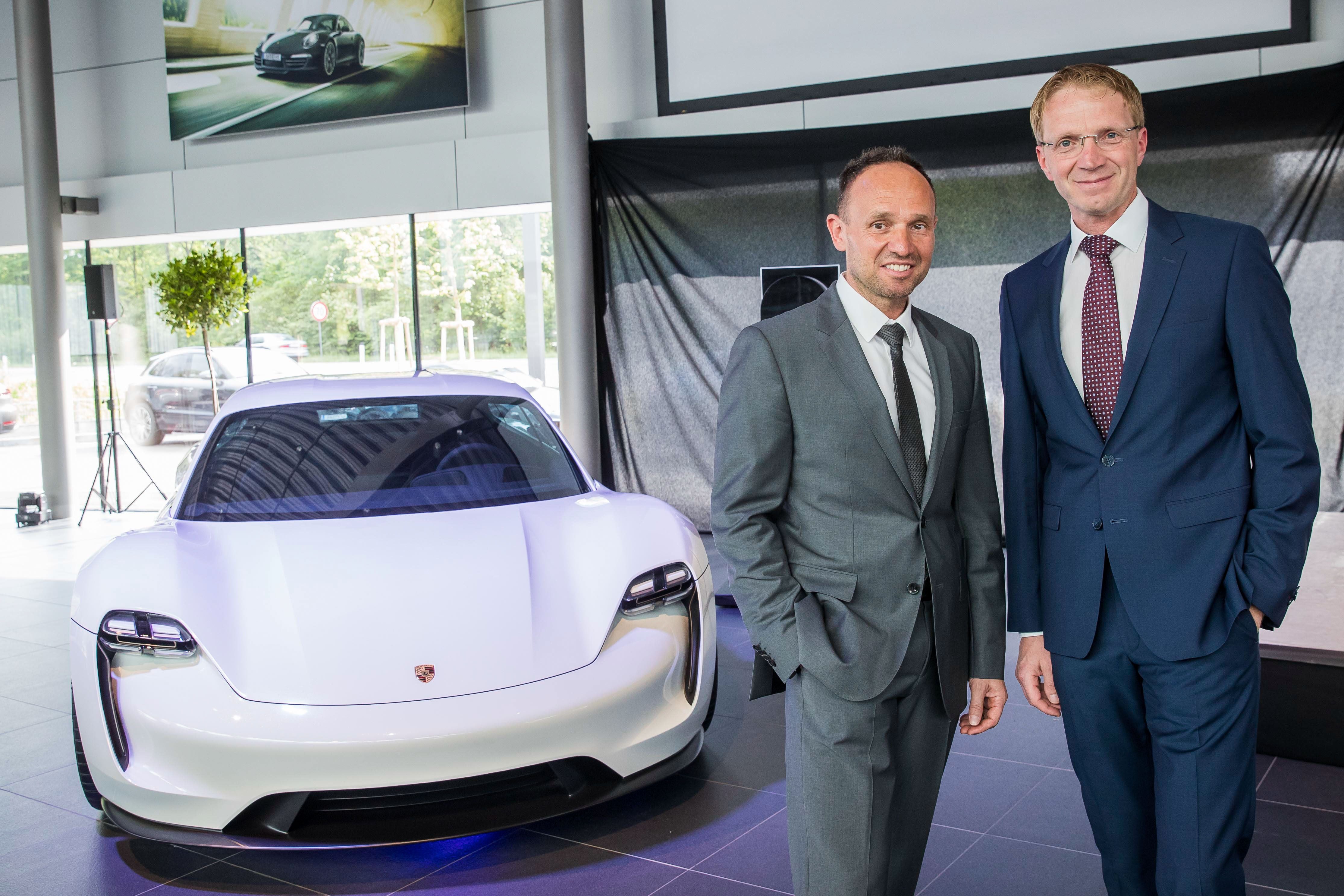Zwei Männer im Anzug stehen vor einem weißen Porsche in einem modernen Autohaus.