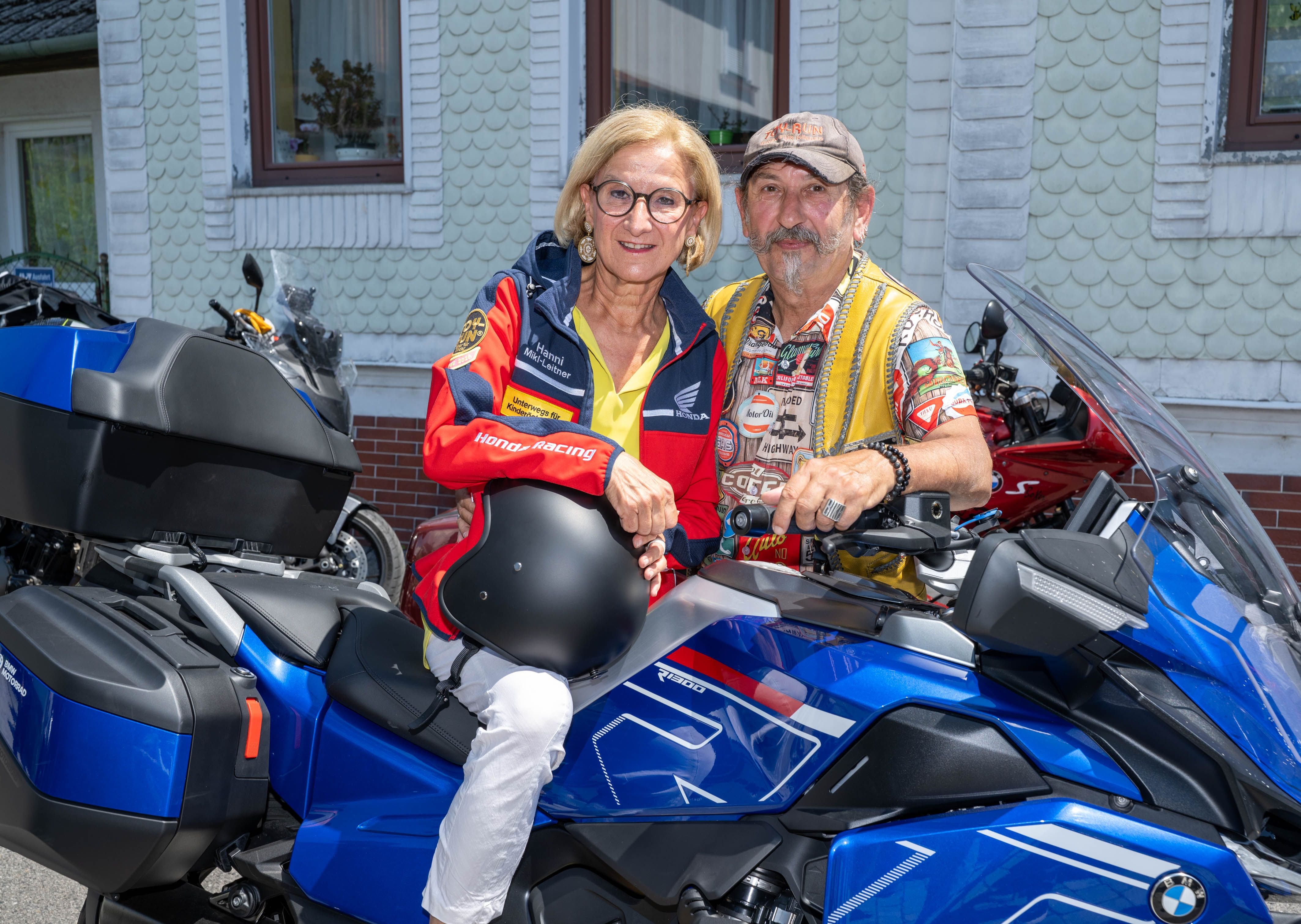 Zwei Personen sitzen auf einem blauen Motorrad vor einem Haus, eine Person hält einen Helm.