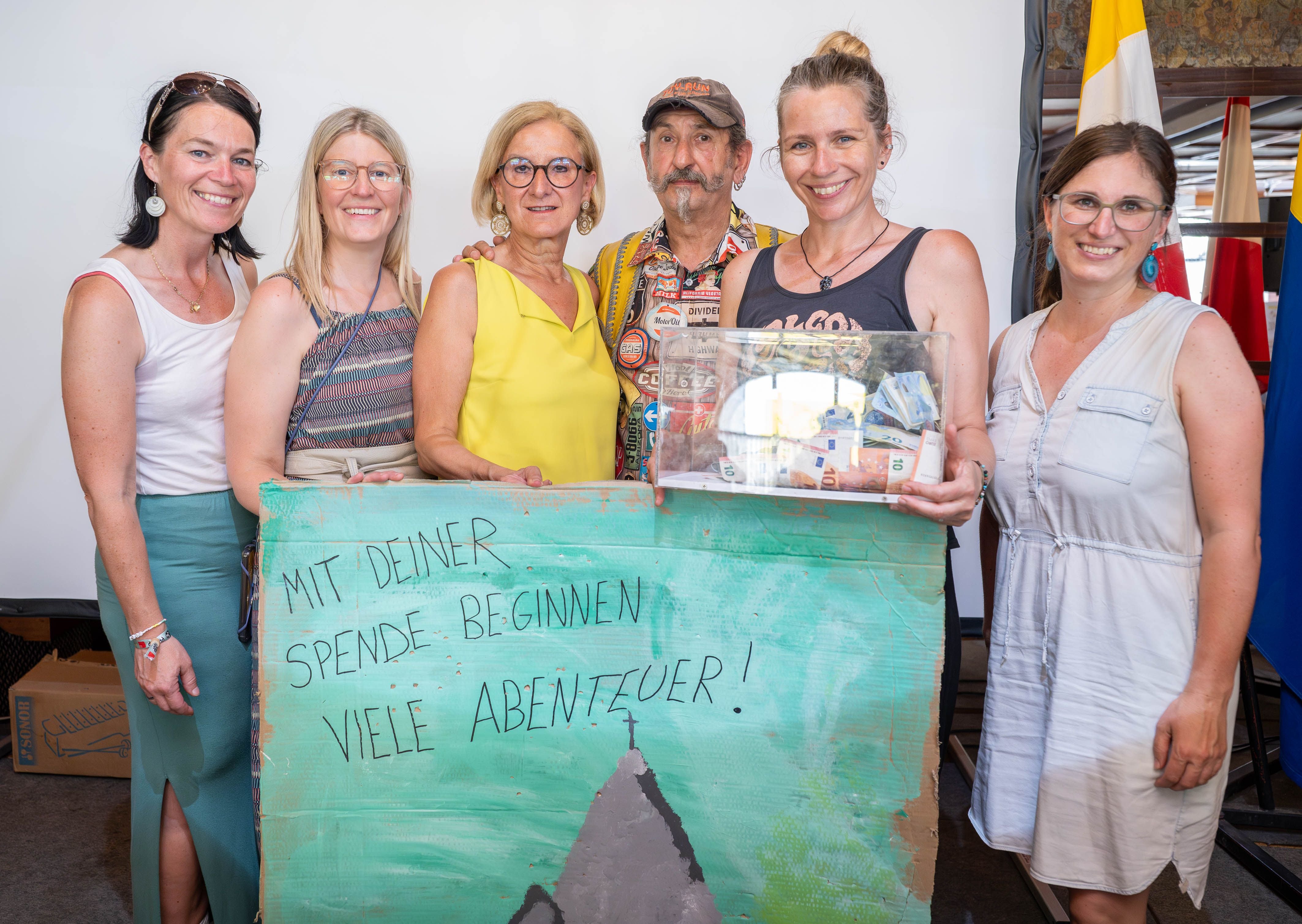 Sechs Personen halten ein Schild mit der Aufschrift „Mit deiner Spende beginnen viele Abenteuer!“ und eine Spendenbox.