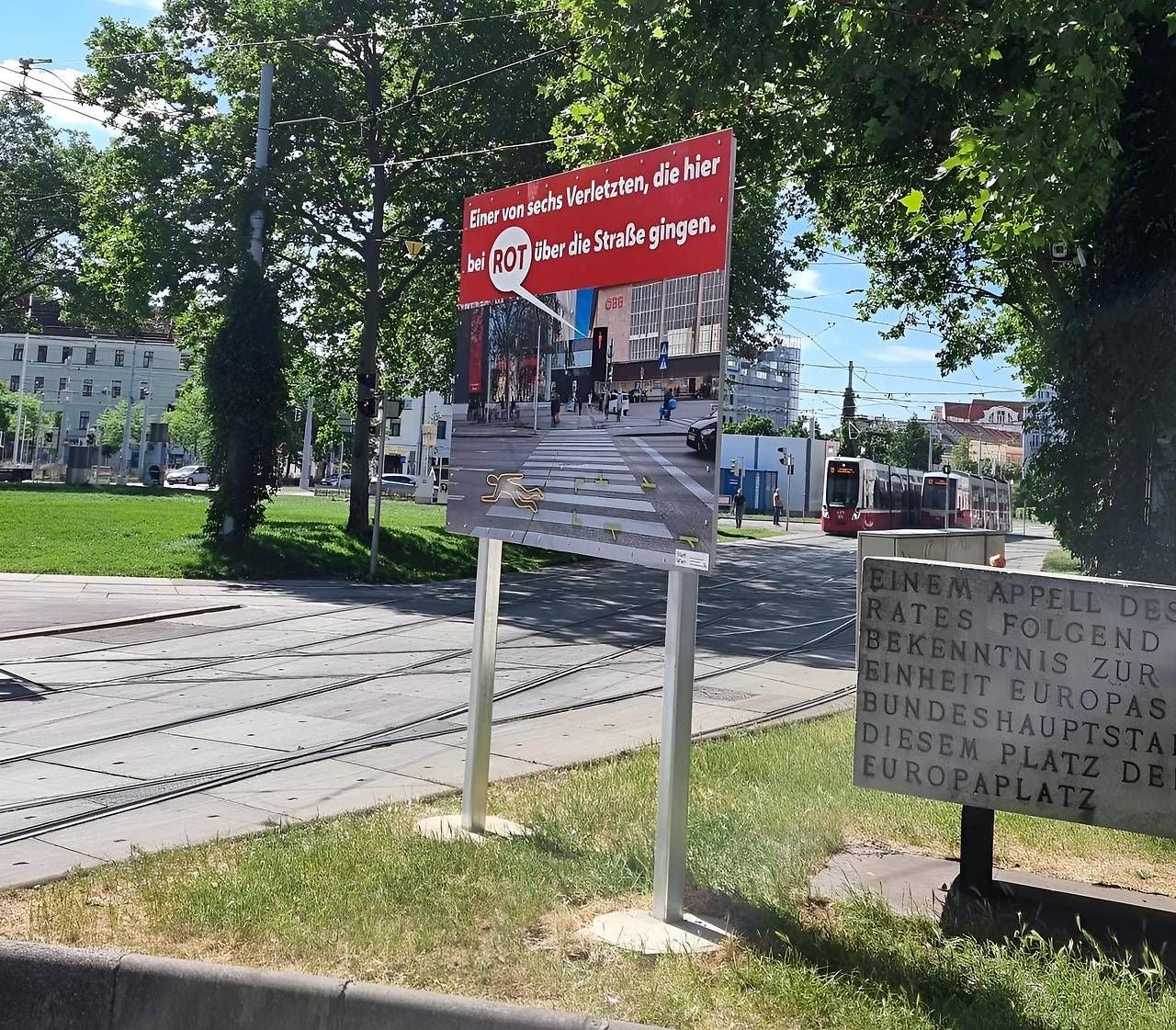 Warnschild warnt Fußgänger davor, bei Rot die Straße zu überqueren, im Hintergrund Straßenbahn.