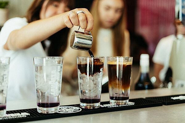 Person gießt Kaffee aus einem kleinen Kännchen in ein Glas mit Eis an einer Bar ein.