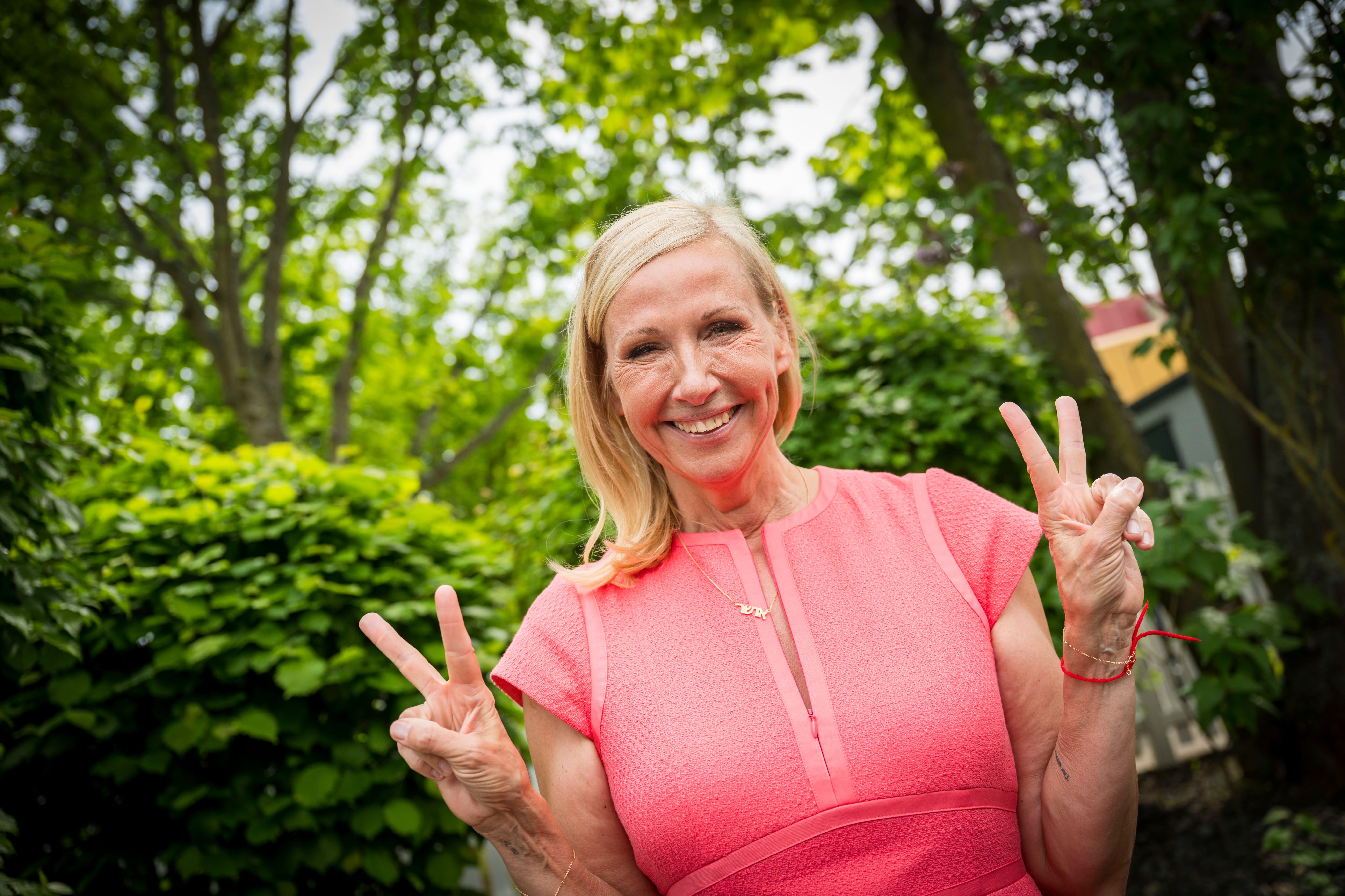 Frau im pinken Kleid zeigt mit beiden Händen das Victory-Zeichen im grünen Garten.