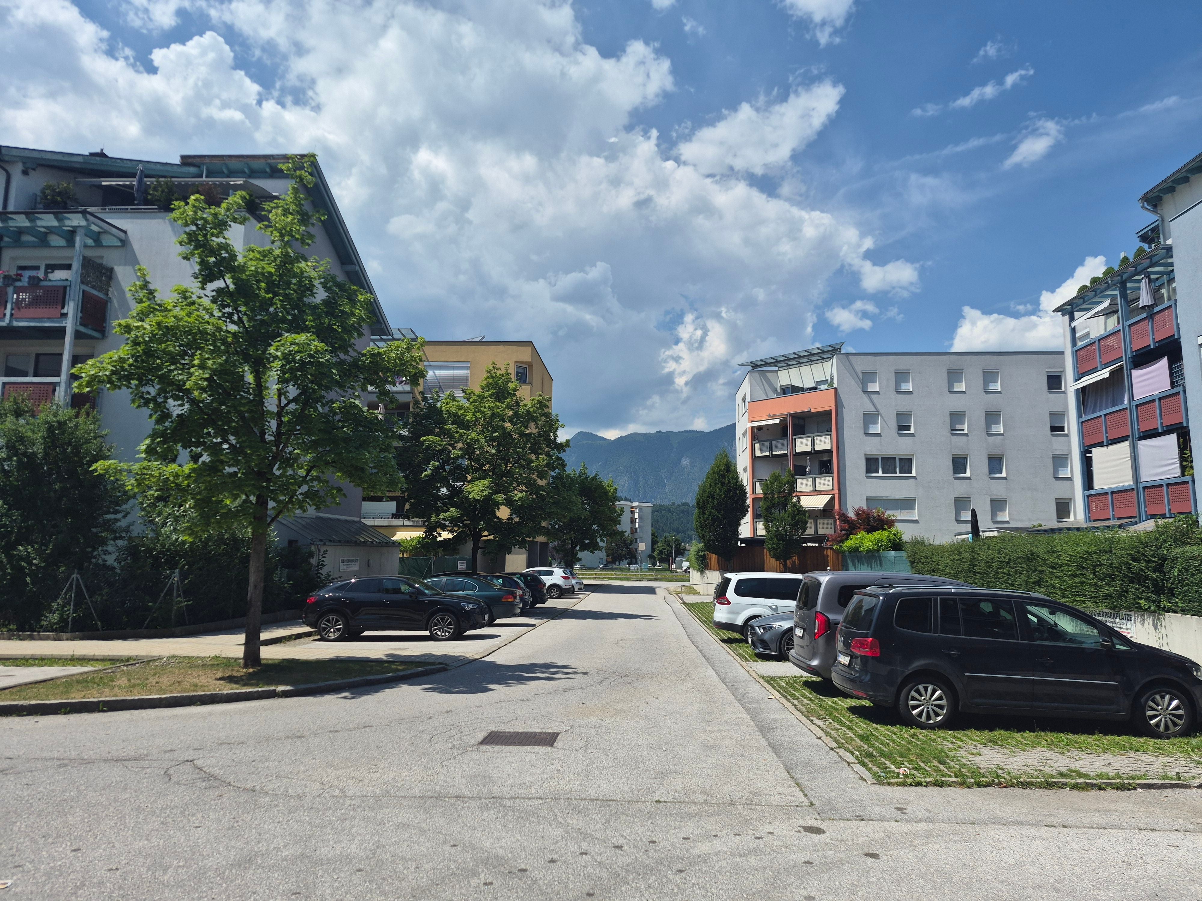 Wohnstraße mit geparkten Autos und Wohnhäusern, im Hintergrund Berge und bewölkter Himmel.