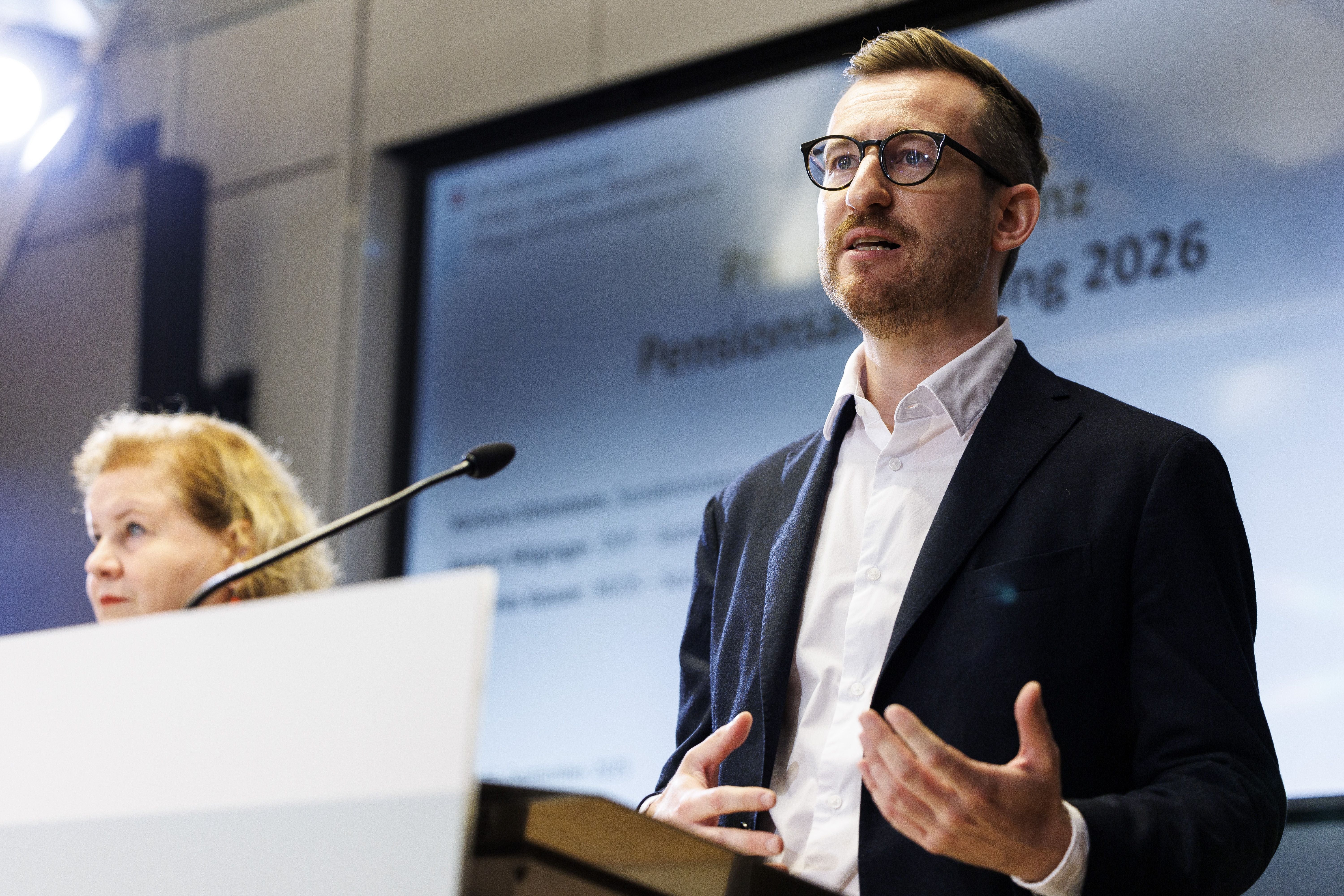 Ein Mann im Anzug spricht bei einer Pressekonferenz, im Hintergrund eine Leinwand.