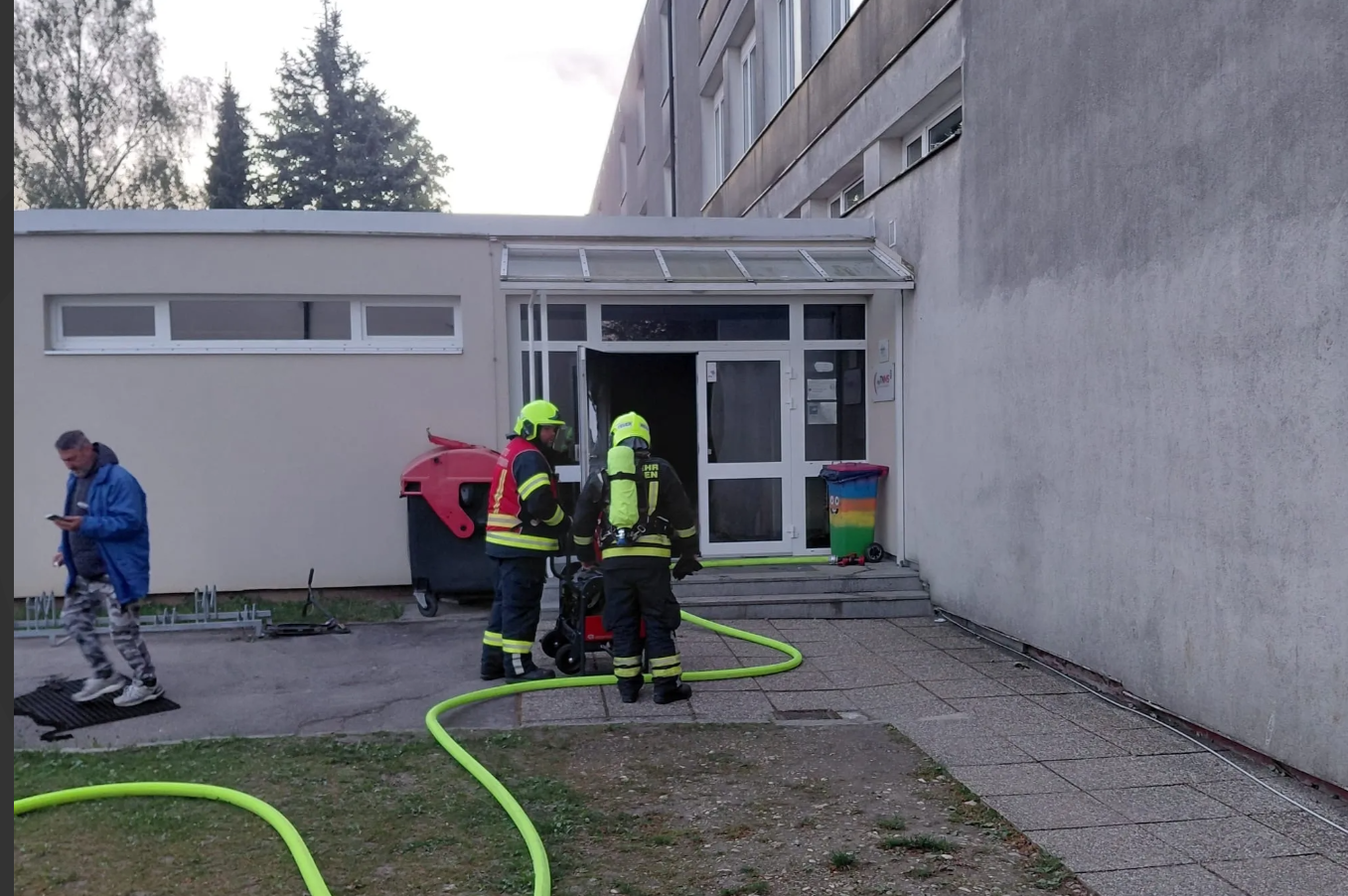 Zwei Feuerwehrleute stehen vor einem verrauchten Eingang, gelbe Schläuche liegen am Boden.
