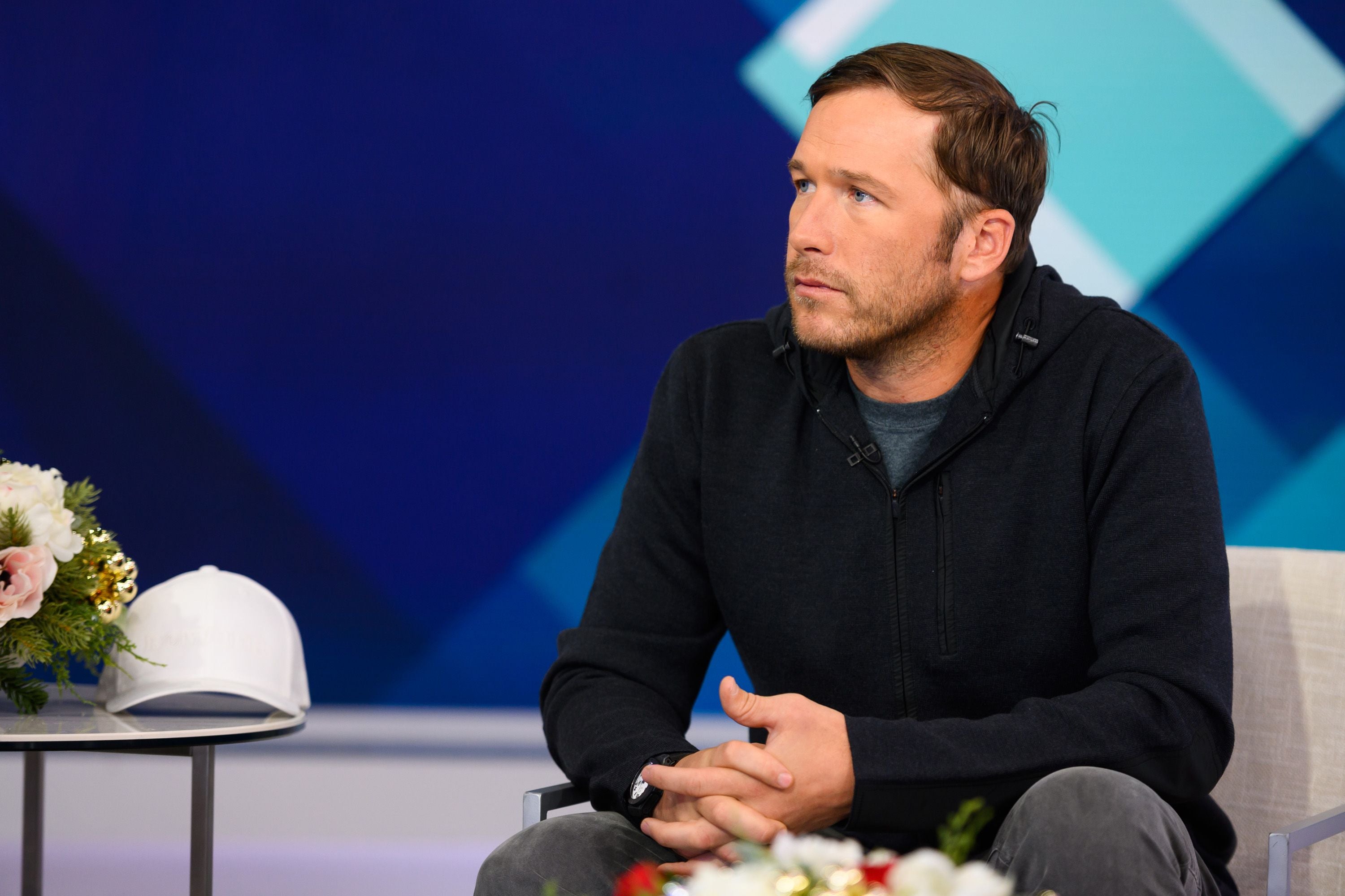 Ein Mann mit dunklem Hoodie sitzt auf einem Stuhl vor blauem Hintergrund.