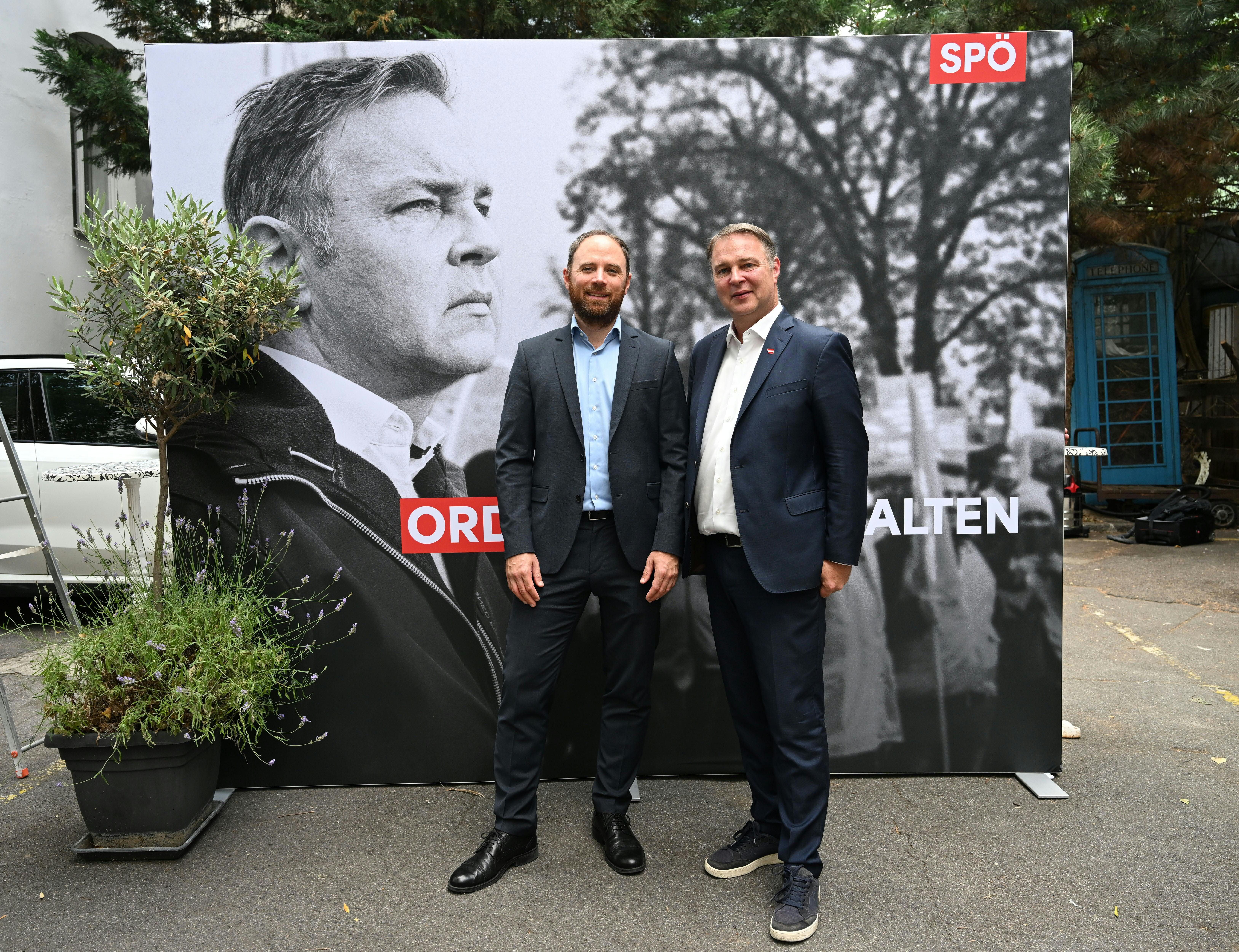 Zwei Männer im Anzug posieren vor einem SPÖ-Plakat bei einer Veranstaltung im Freien.