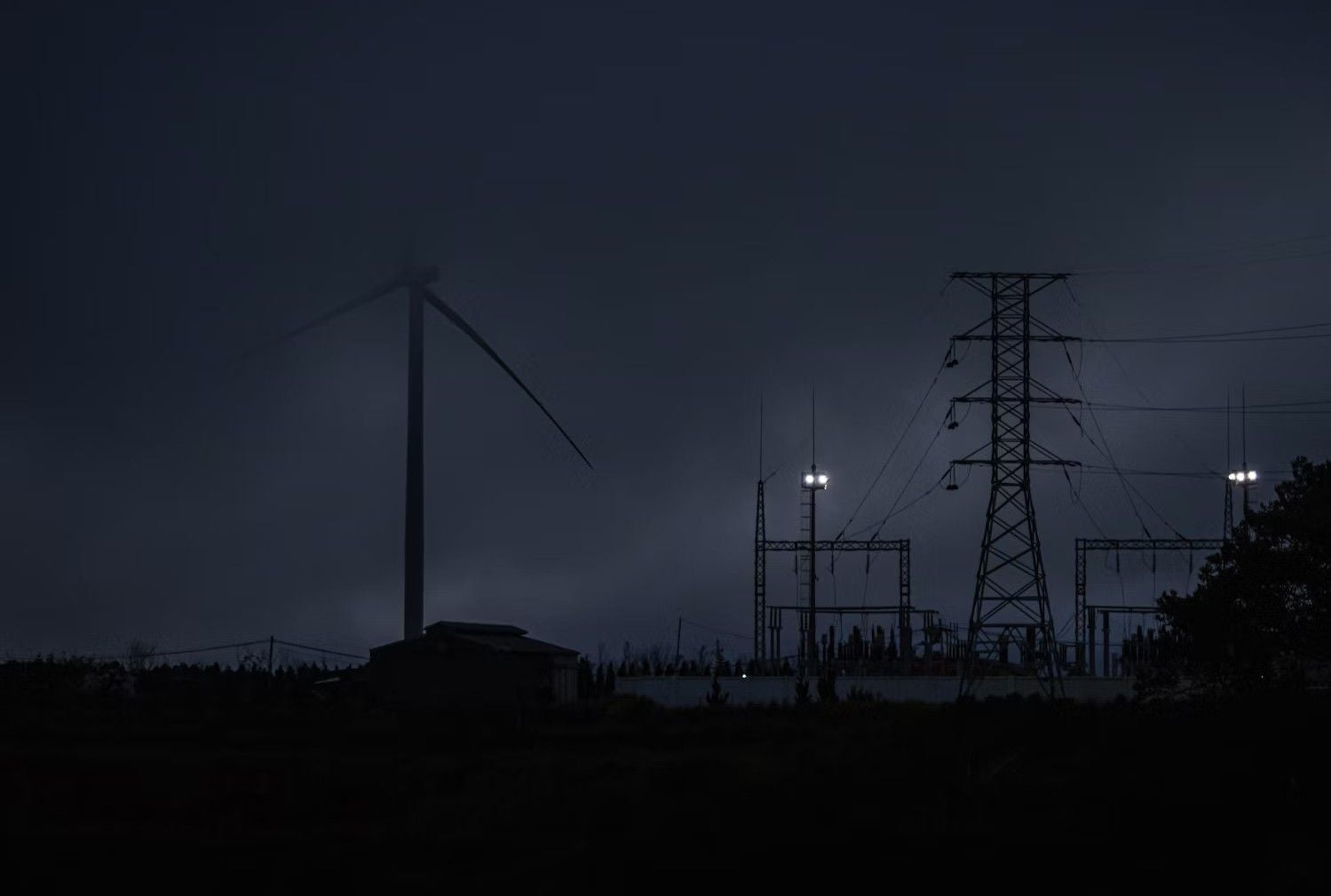 Windrad und Strommast bei Nacht, einige Scheinwerfer beleuchten eine Umspannstation.