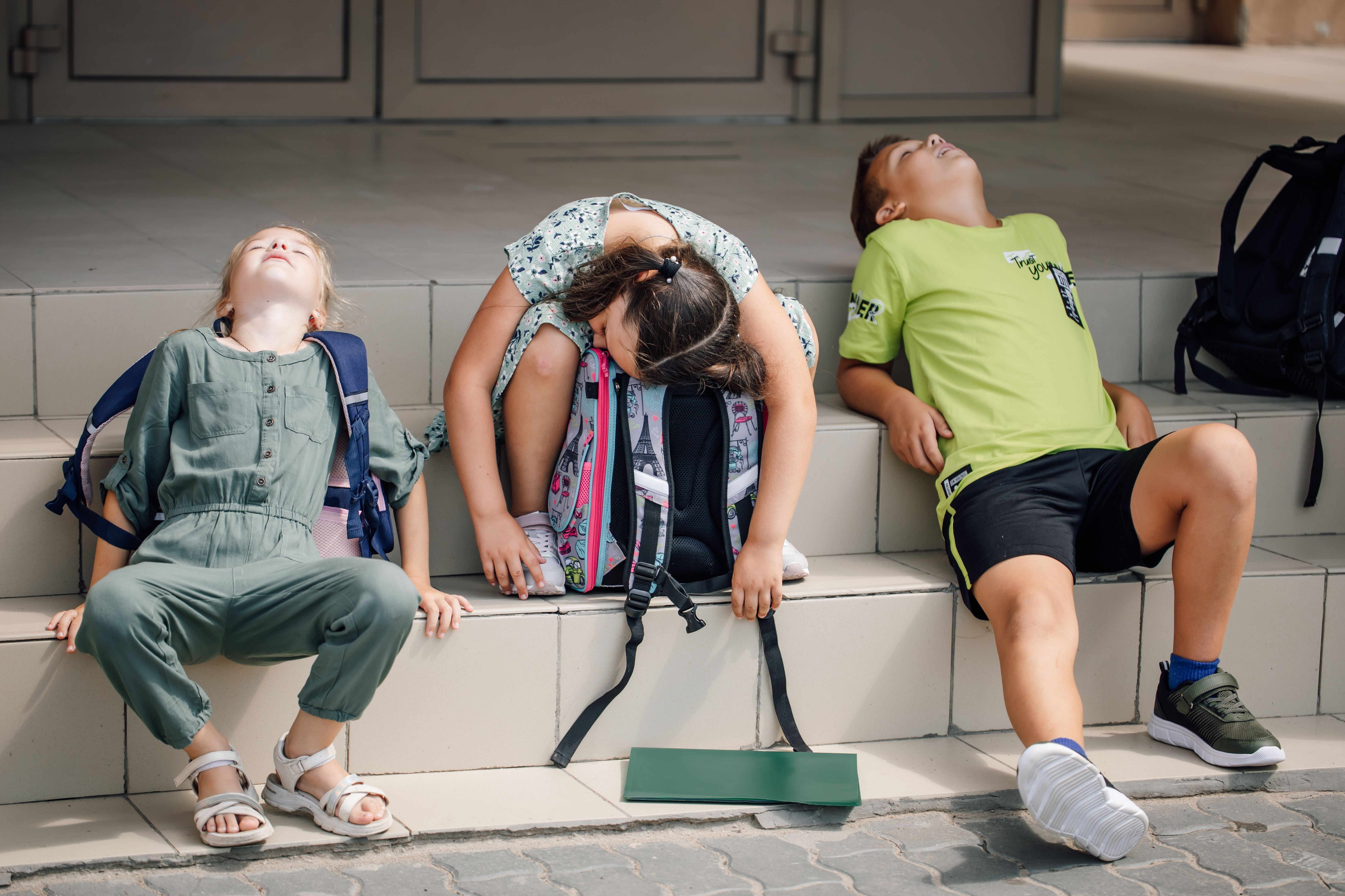 Drei erschöpfte Schulkinder sitzen entspannt mit Rucksäcken auf Treppenstufen.