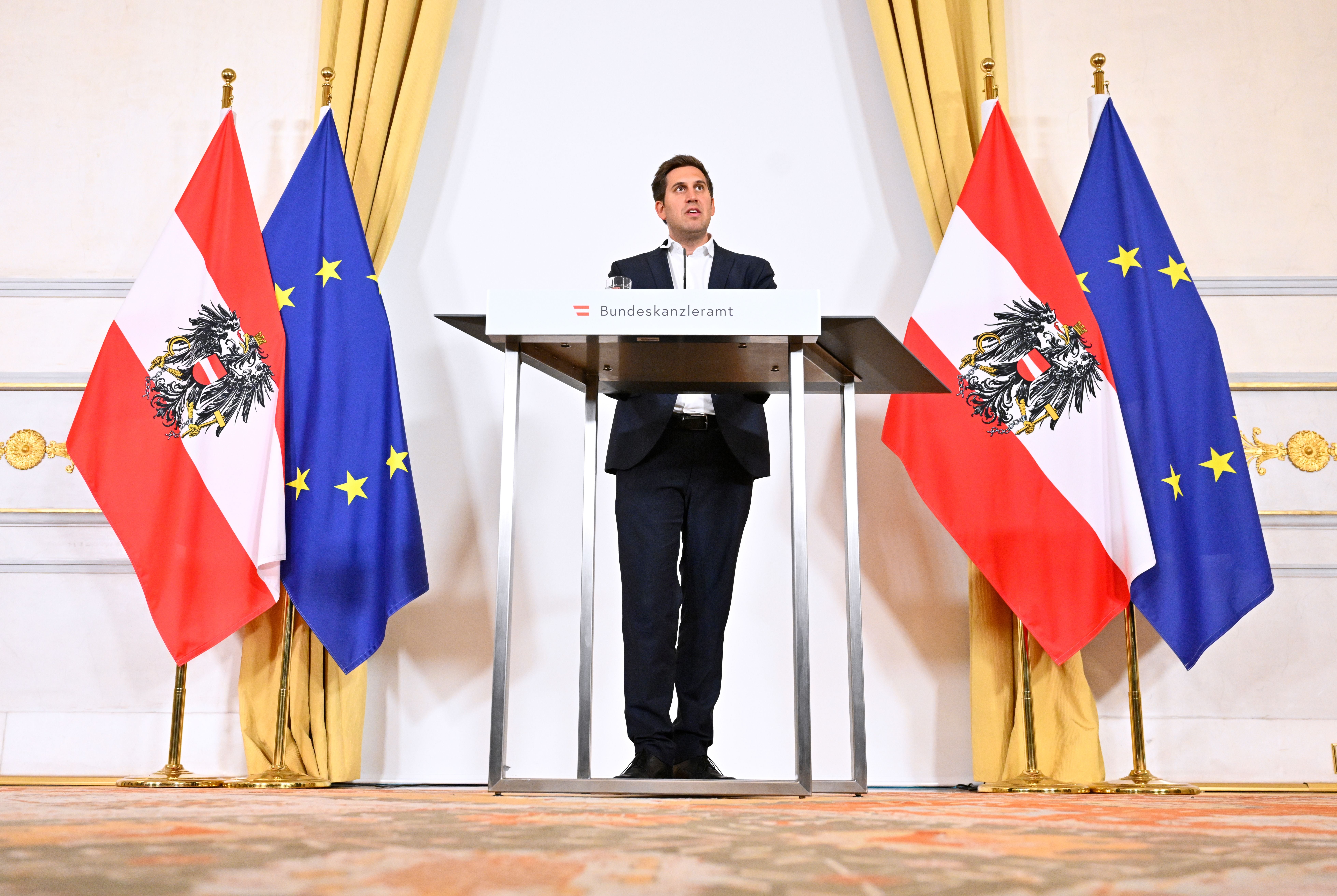 Ein Mann steht am Rednerpult im Bundeskanzleramt, flankiert von Österreich- und EU-Flaggen.