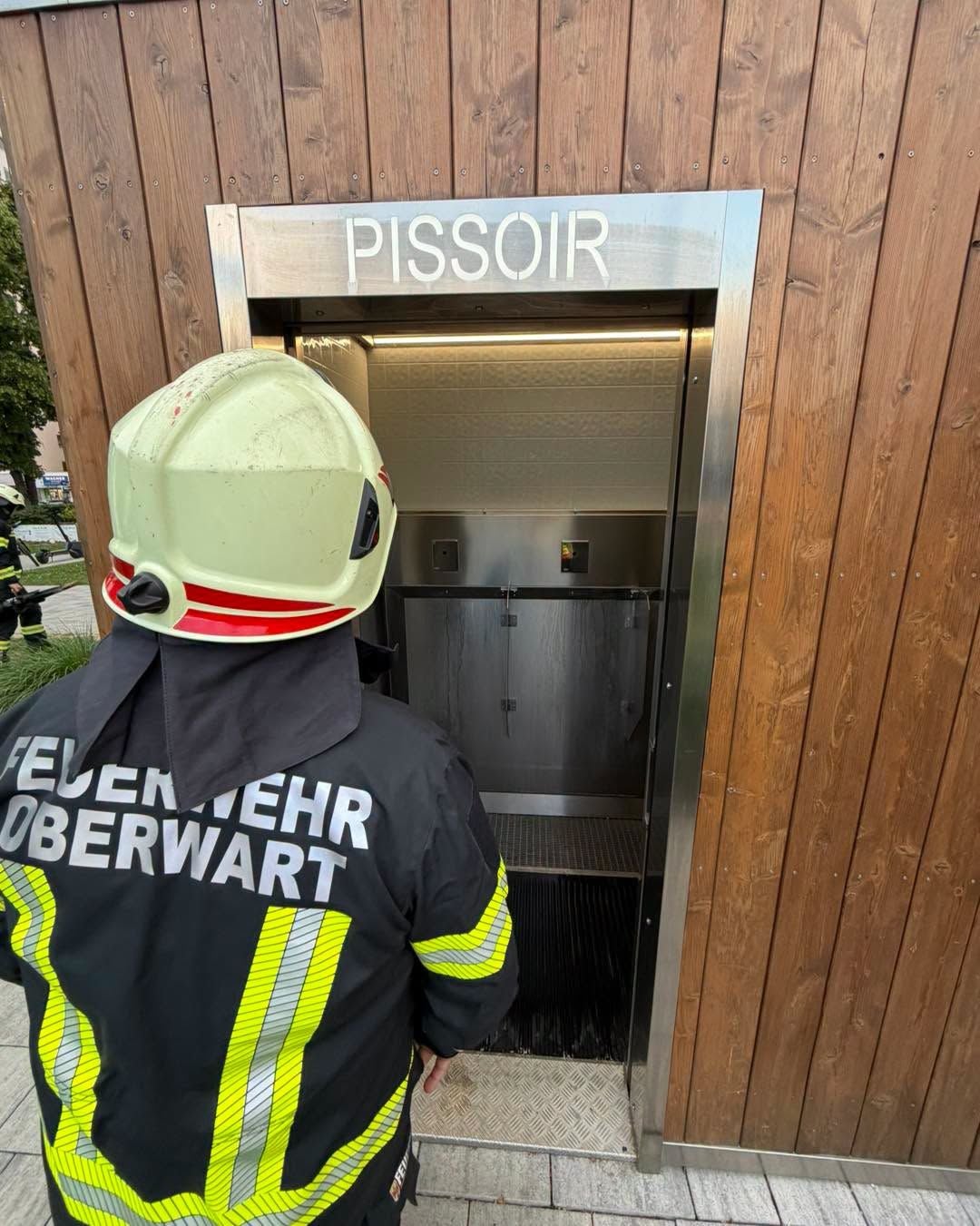 Feuerwehrmann steht vor einem öffentlichen Pissoir mit Holzverkleidung.