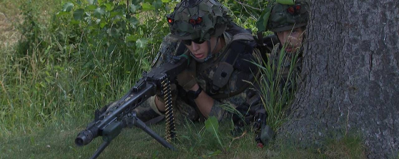 Zwei Soldaten in Tarnuniform verstecken sich mit Maschinengewehr hinter einem Baum im Gras.